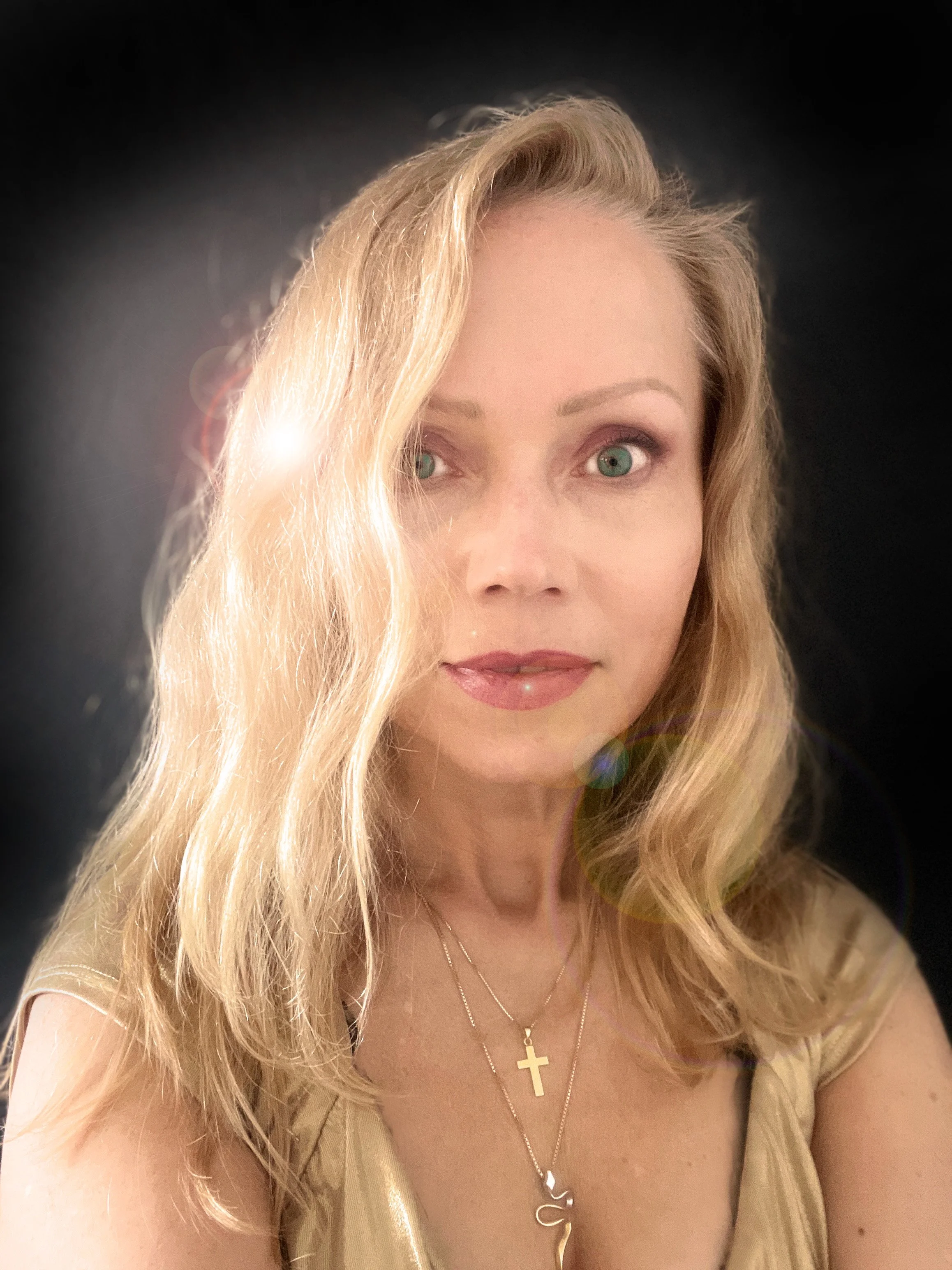 A woman with long blonde hair, blue eyes, wearing a beige top, gold necklaces including a cross, and a pendant with the letter 'J'. The background is black.