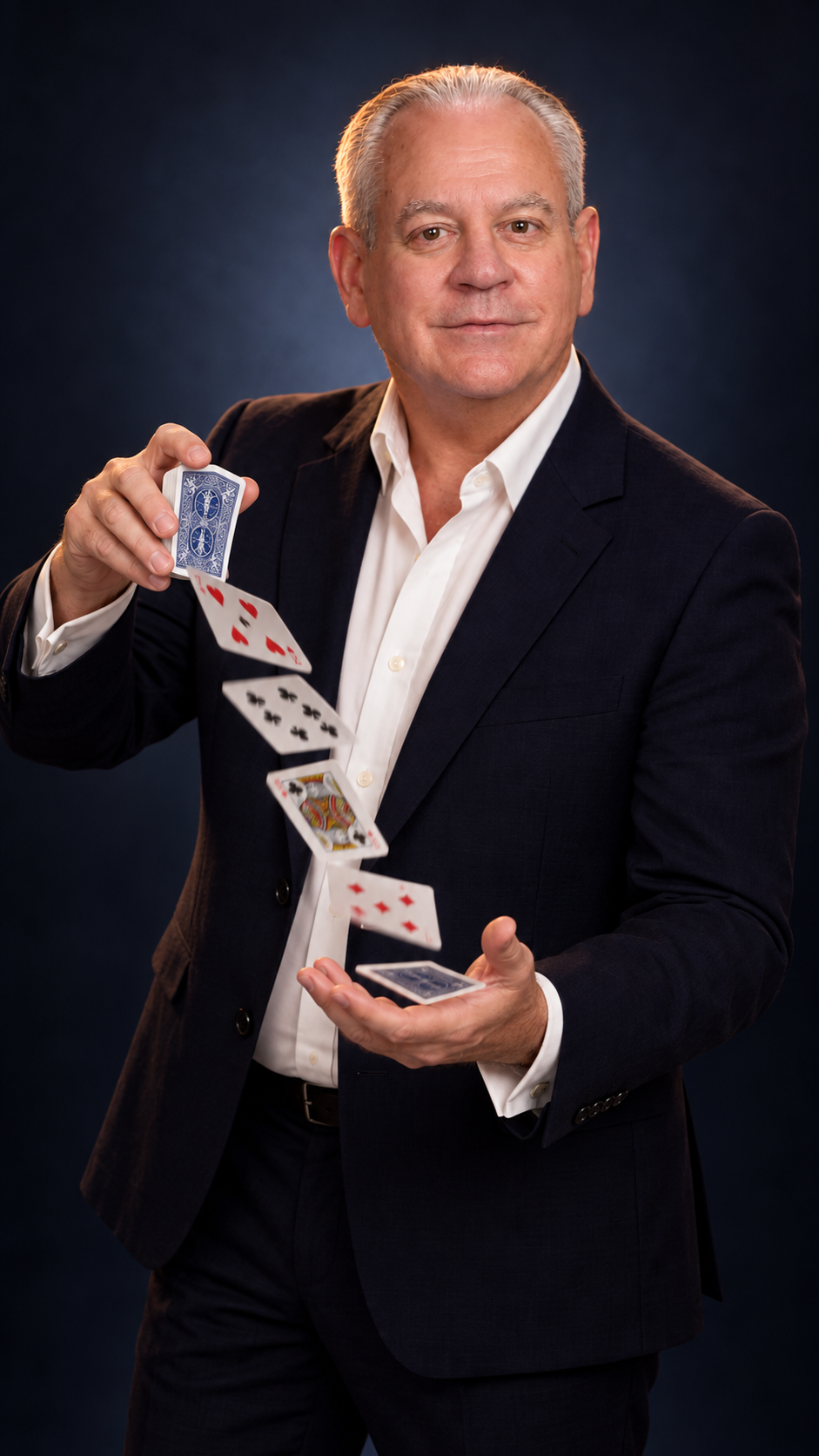 A man in a black suit and white shirt performing a card trick with playing cards, with cards floating in midair against a dark background.