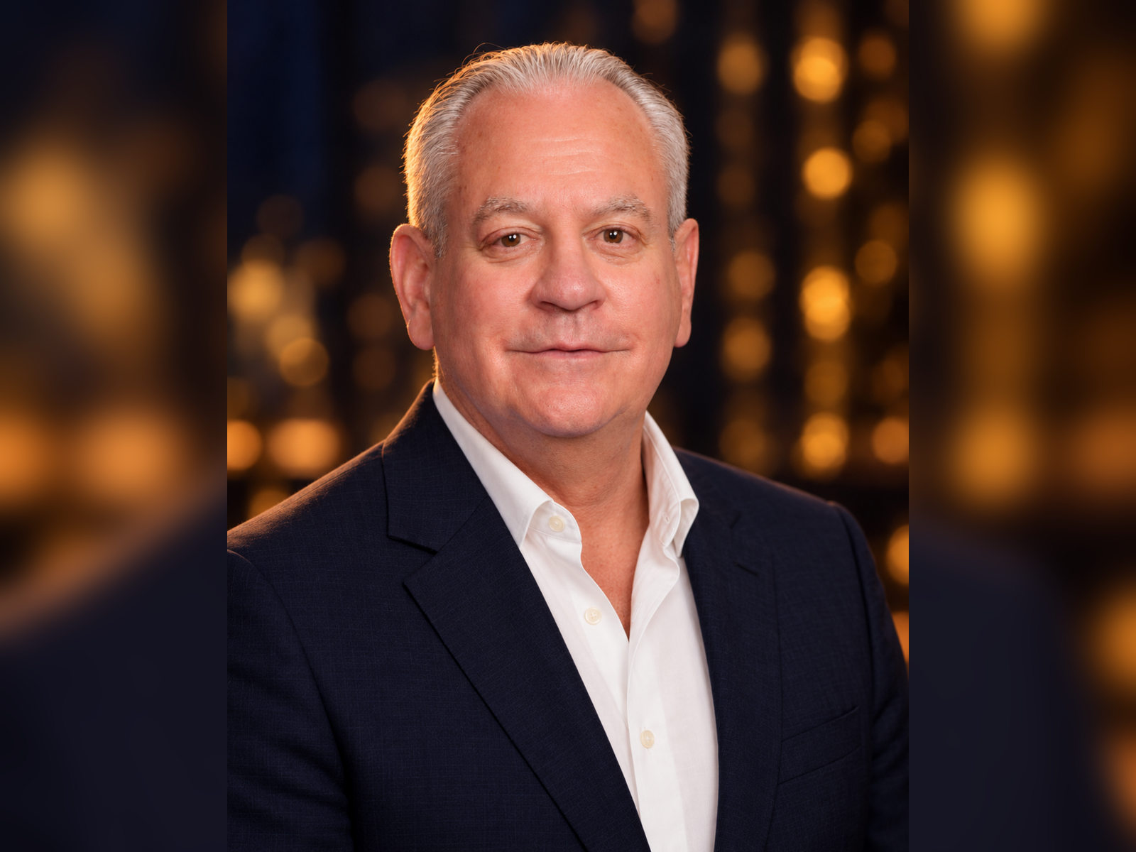 A middle-aged man with short gray hair wearing a white shirt and dark blazer, posing for a portrait against a warm, out-of-focus background with golden lights.