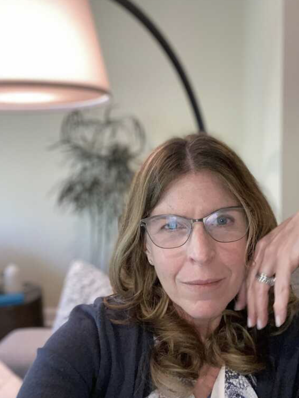 A woman with brown hair and glasses smiling, sitting indoors with a lamp and a decorative plant in the background.