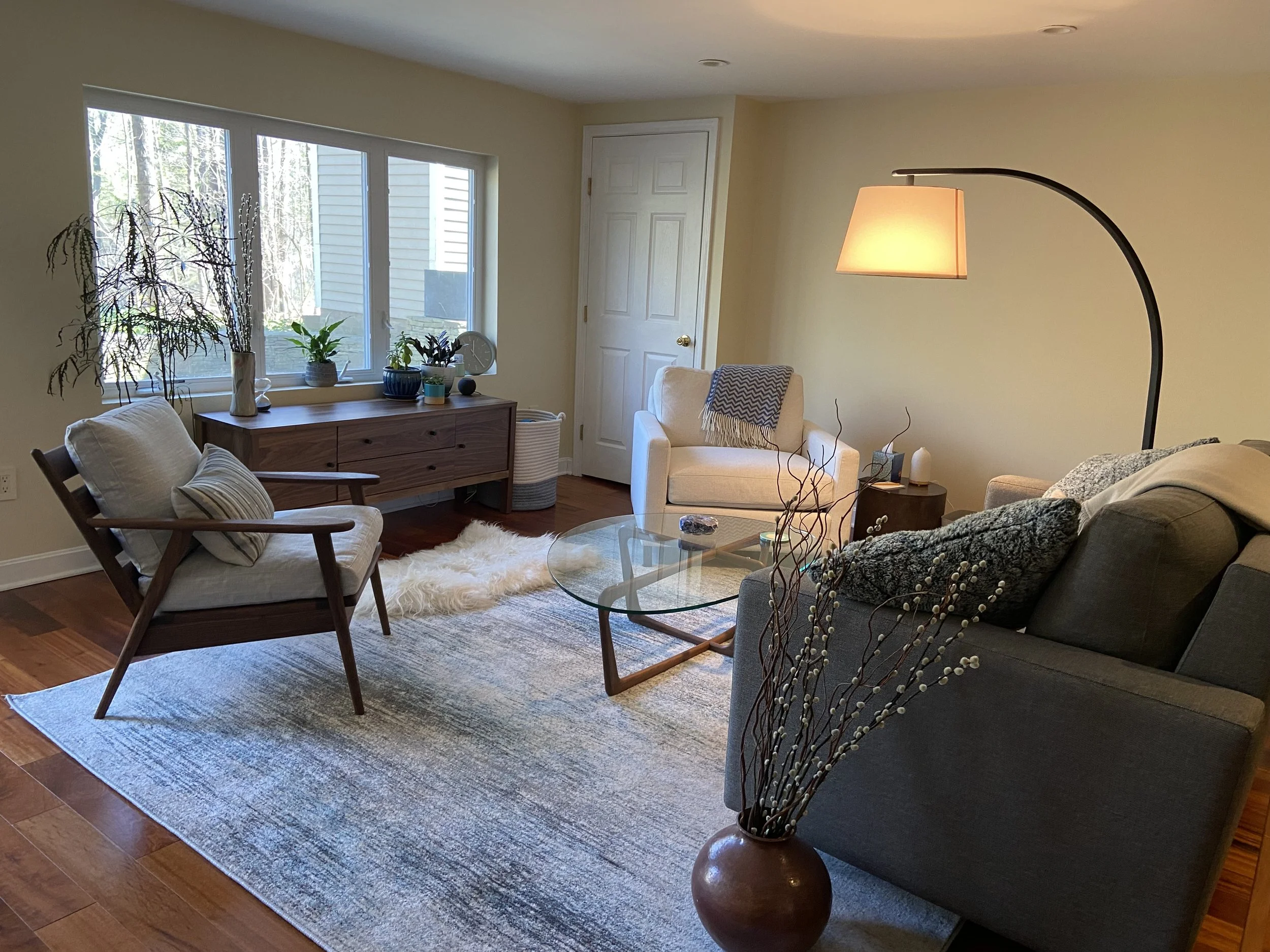 A cozy living room with a window showing trees outside. There is a wooden sideboard with potted plants, a white armchair with a patterned throw, a gray couch with a blanket, a glass coffee table, a fluffy rug, and a floor lamp with a curved stand.