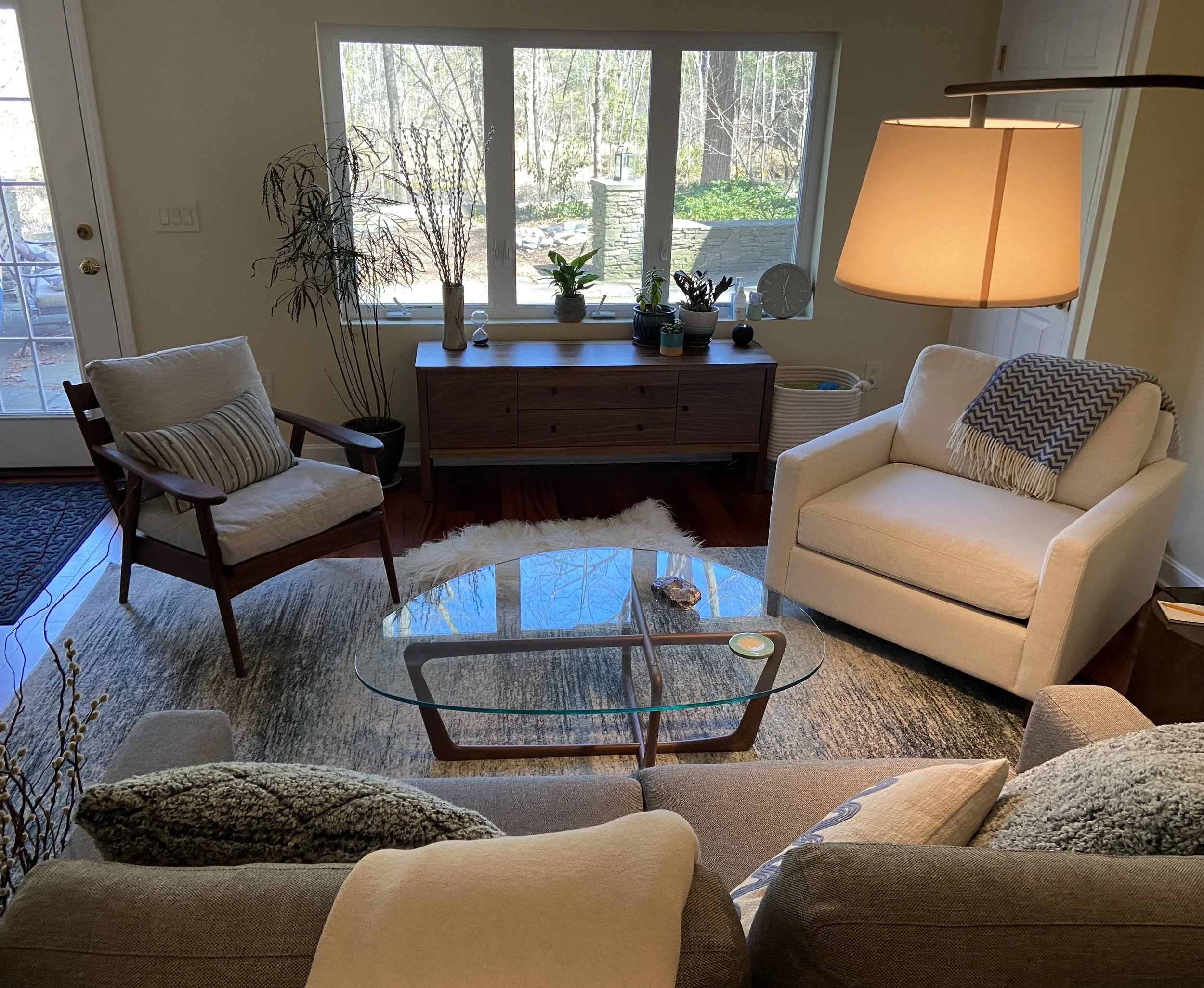 Living room with beige sofas, a glass coffee table, a wooden sideboard with potted plants, a large window with outdoor trees, and a hanging lamp.