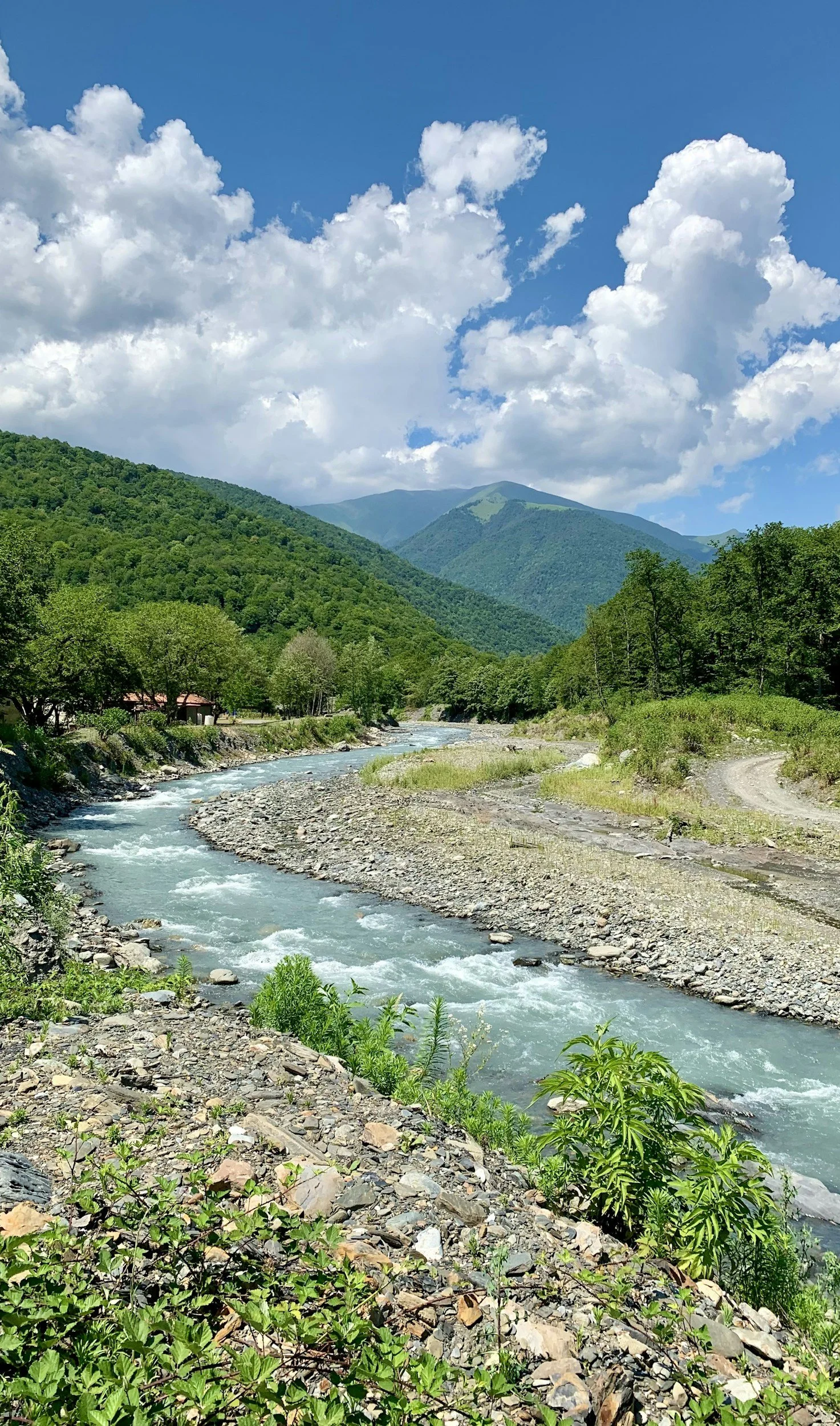 A mountain landscape with green hills and a flowing river in the foreground, under a partly cloudy blue sky.