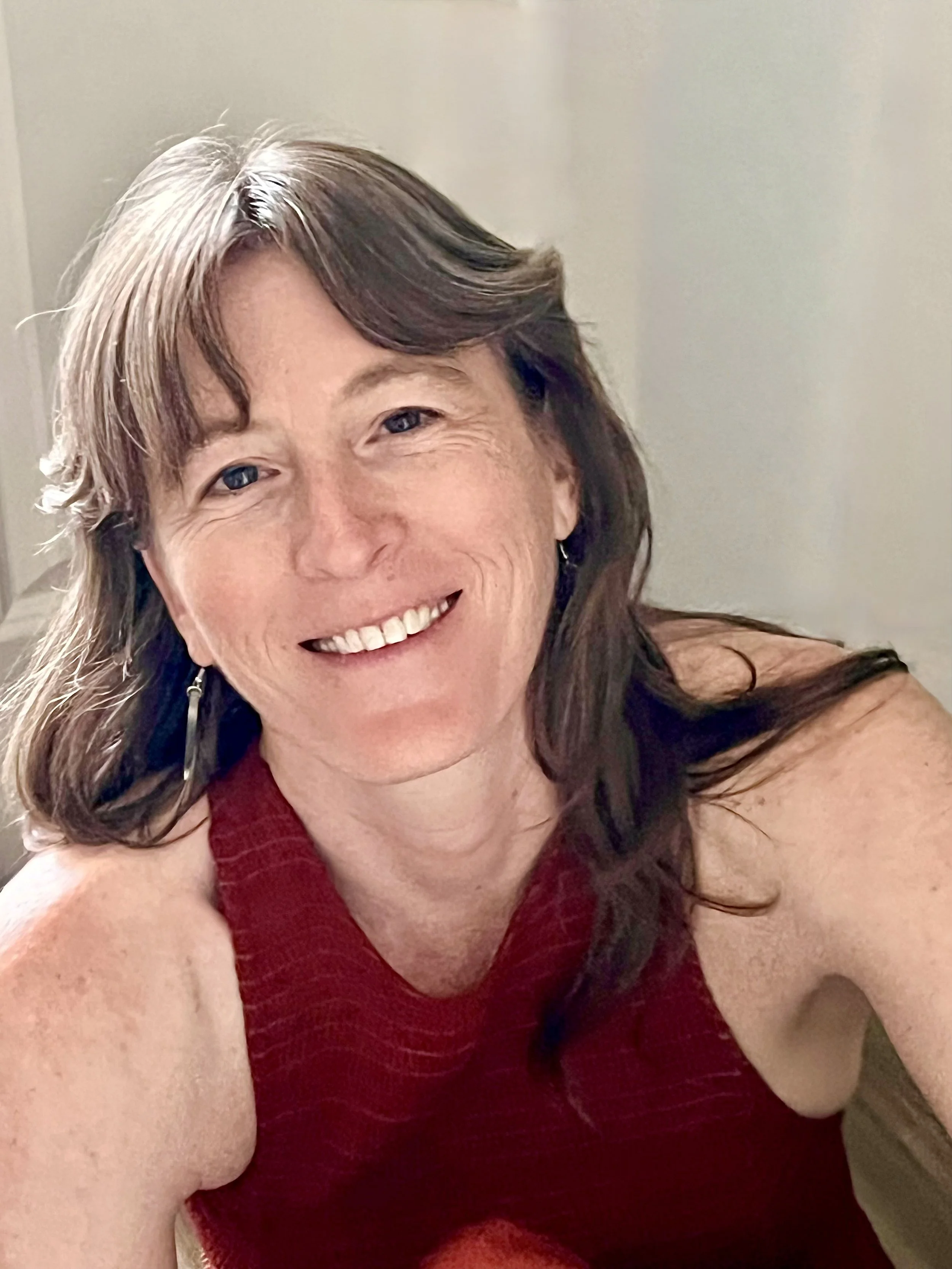 A smiling woman with shoulder-length brown hair, blue eyes, and wearing a sleeveless red top, taking a selfie in a well-lit room.