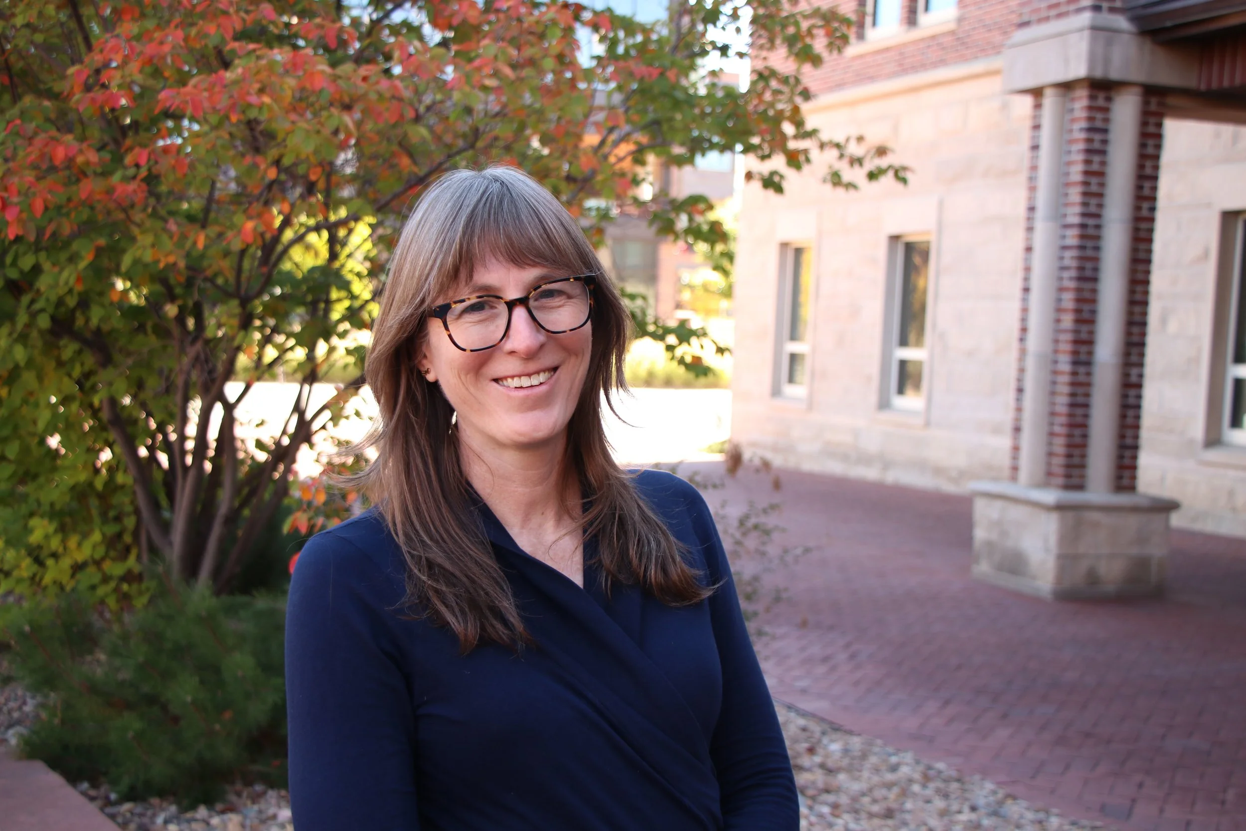 A woman with long brown hair, wearing glasses and a navy blue top, smiling outdoors in front of a red brick building and a tree with amber and green leaves.