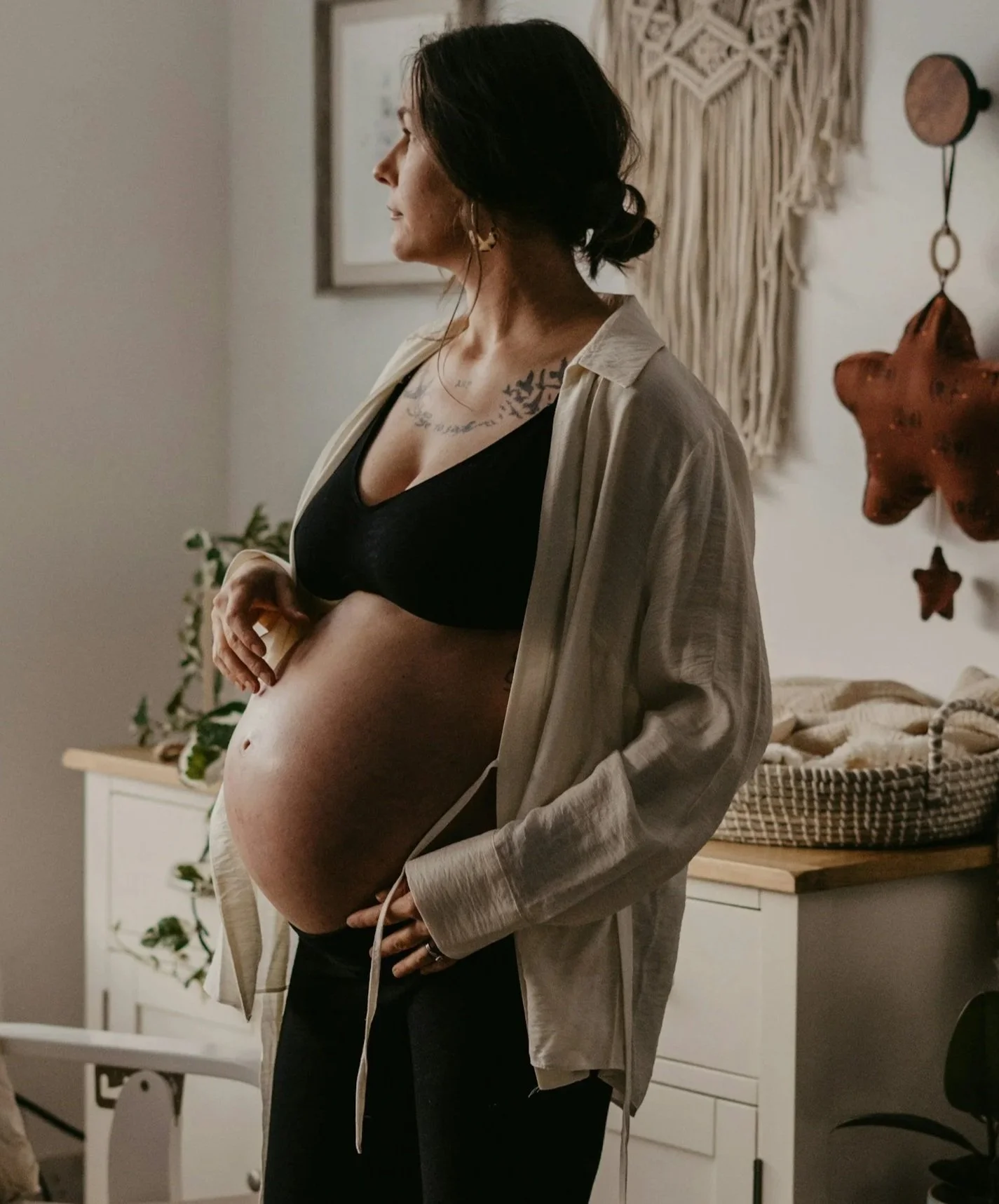 Pregnant woman in black bra and beige shirt, standing sideways in a cozy, decorated room, with wall art and a star-shaped ornament in the background.