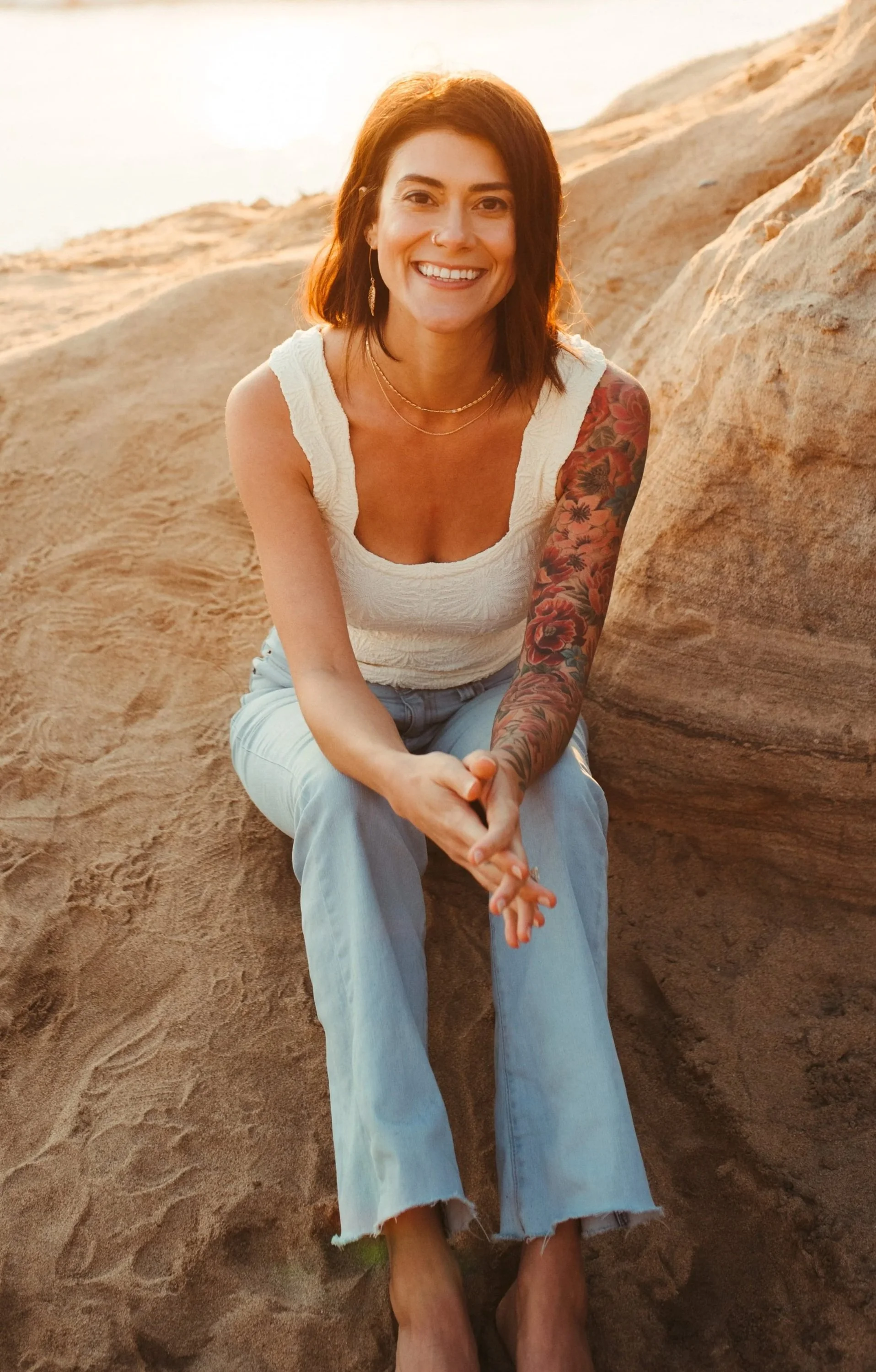 A woman with red hair and tattoos on her left arm, smiling while sitting on a rock in a desert-like landscape during sunset.