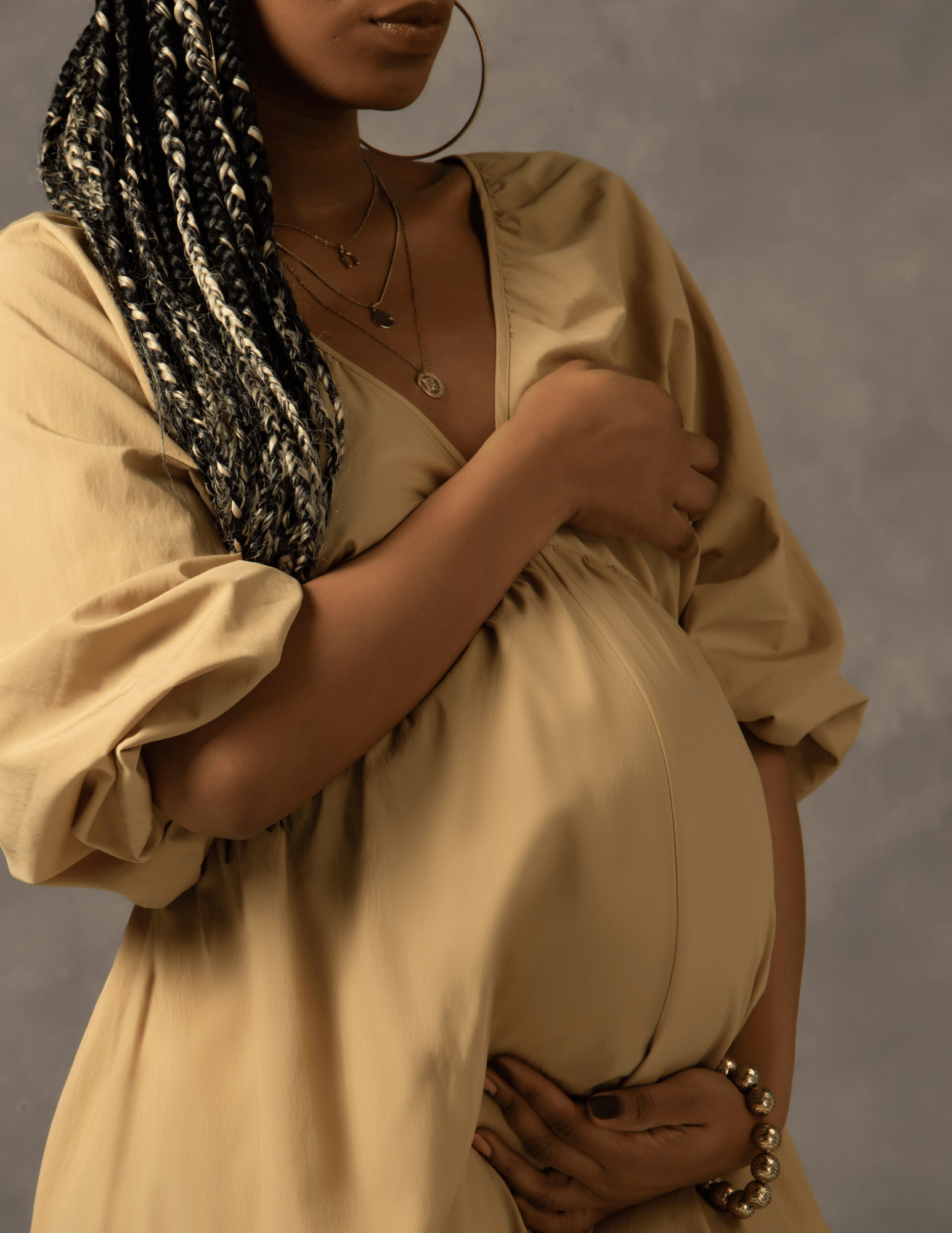 A pregnant woman wearing a beige dress with puffed sleeves, layered necklaces, hoop earrings, and a beaded bracelet, standing against a neutral background.