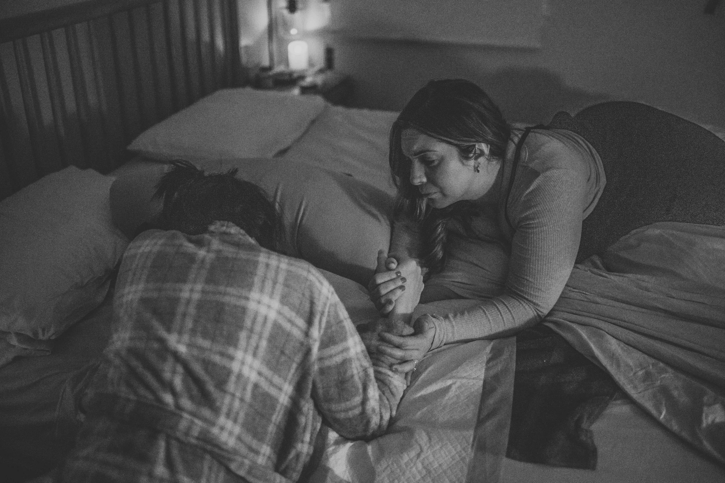 A woman kneeling on a bed, looking at a person lying down with their face turned away. The scene appears to be at night, with dim lighting and a bedside lamp on.