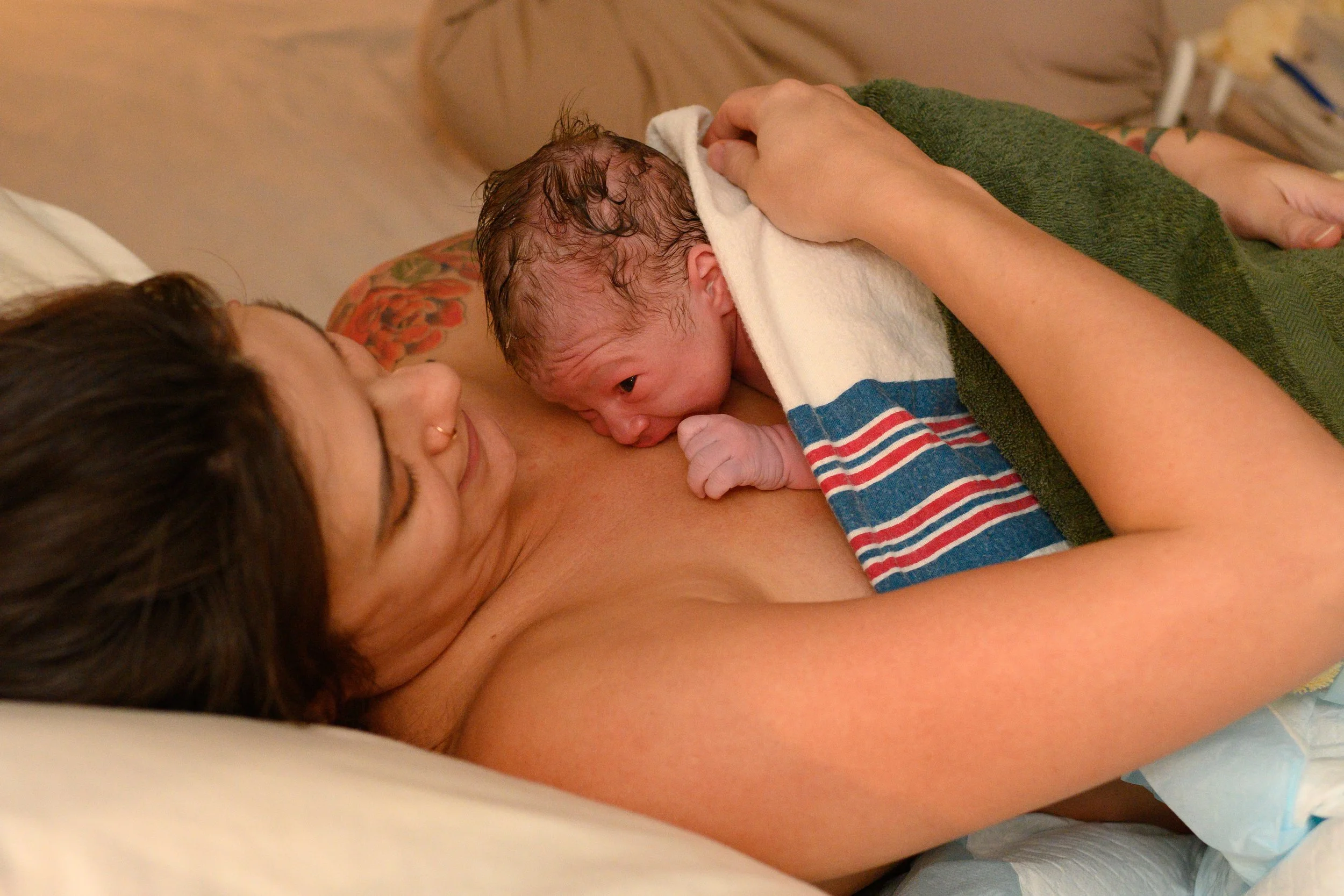 A newborn baby being gently wiped with a white towel by a woman, lying on a bed with a tattoo on her shoulder and a green towel covering the baby's body.