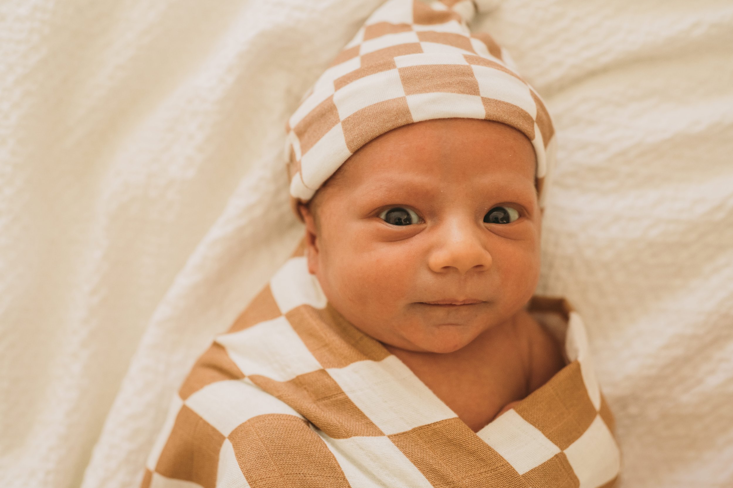 Close-up of a baby with a checkered hat and matching outfit, lying on a soft surface, looking at the camera.