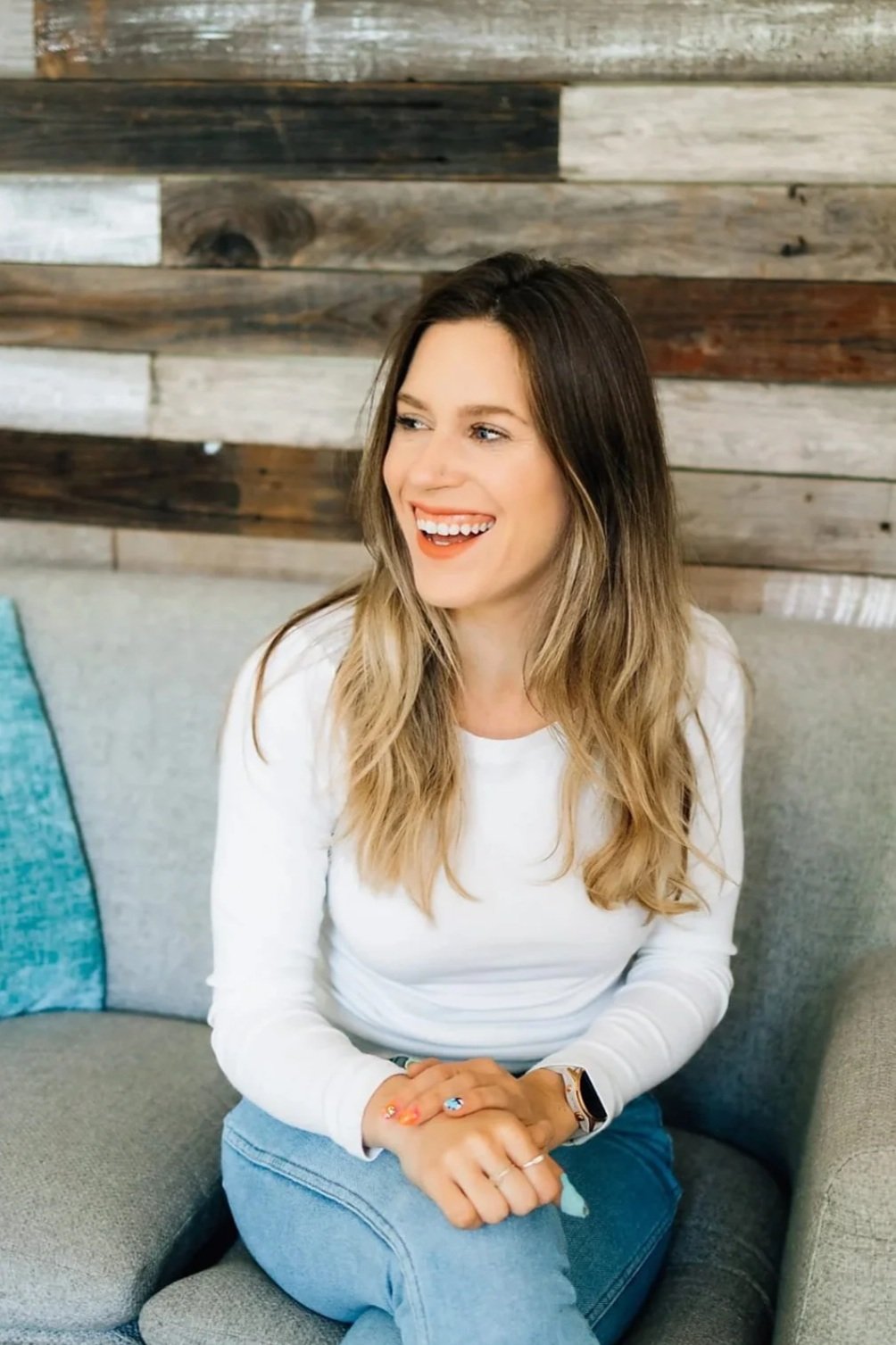 A woman with long, wavy blonde hair and fair skin, wearing a white shirt and blue jeans, sitting on a grey couch with a rustic wood-paneled wall behind her, smiling and looking to her right.