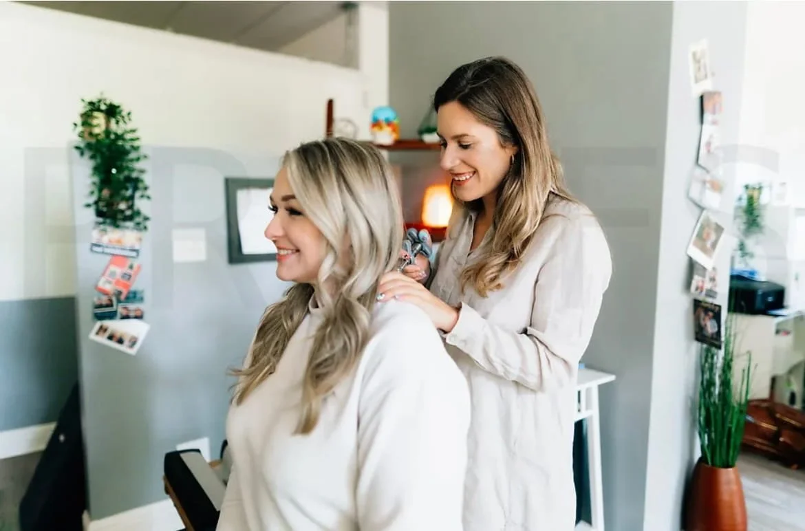 Two women in a cozy living room, one getting her hair styled by the other, both smiling.
