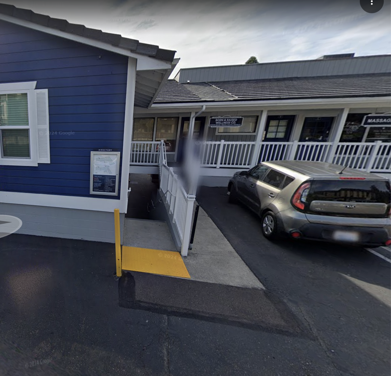 View of a blue building with a white sign and an adjacent gray building with signs for wellness and massage services, a car parked in front, and a small ramp with a yellow safety barrier.