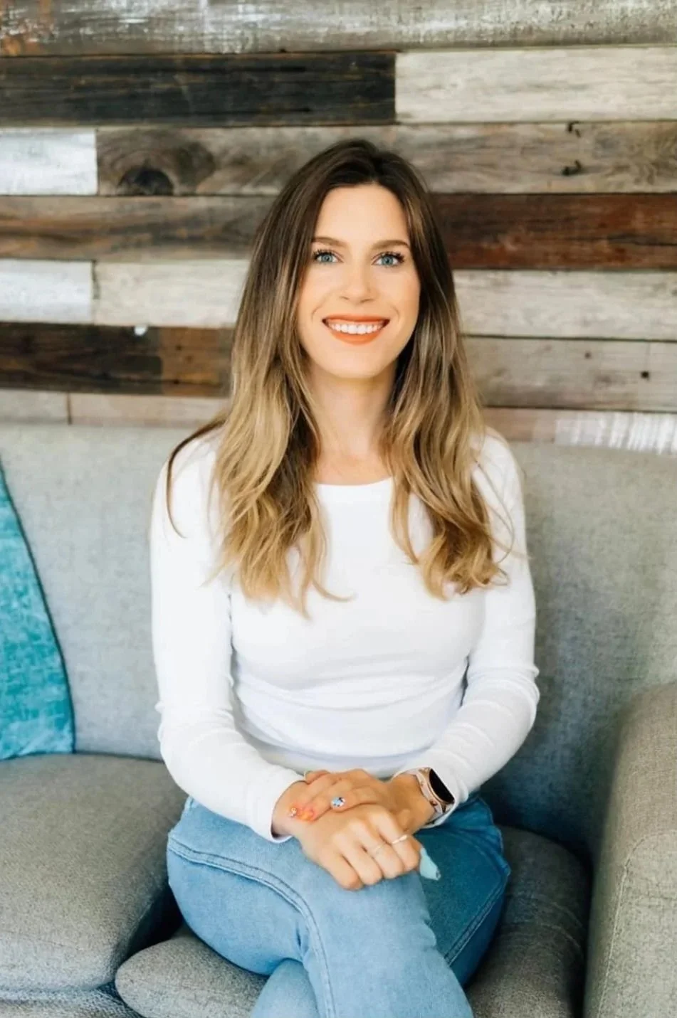 A smiling woman with long brown hair, wearing a beige and white striped shirt and leopard-print pants, sitting with her arms wrapped around her knees against a plain beige wall.