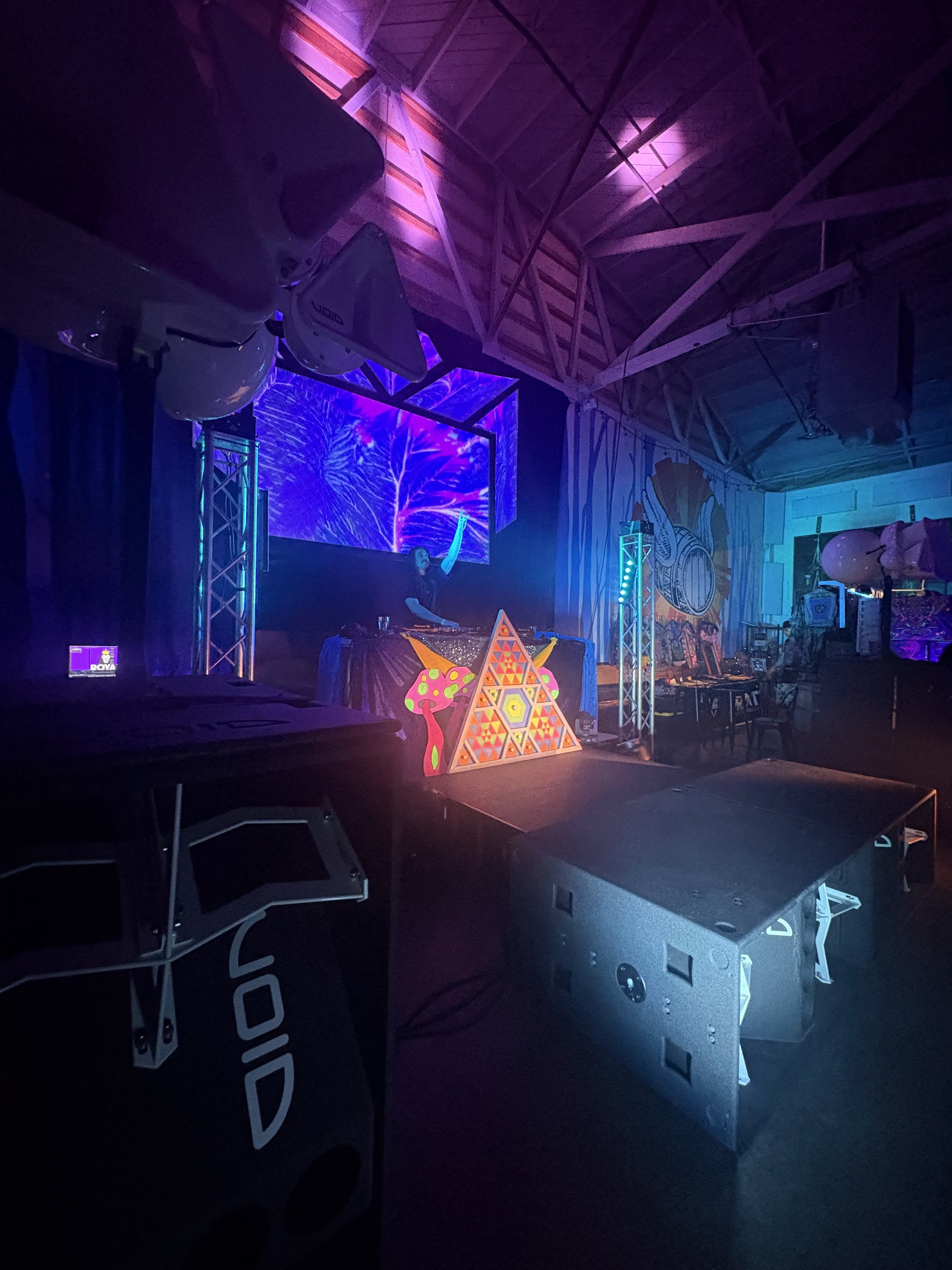 An indoor dance music event with DJ equipment, colorful psychedelic designs, a digital screen displaying visual effects, and decorative neon-lit elements on the stage.