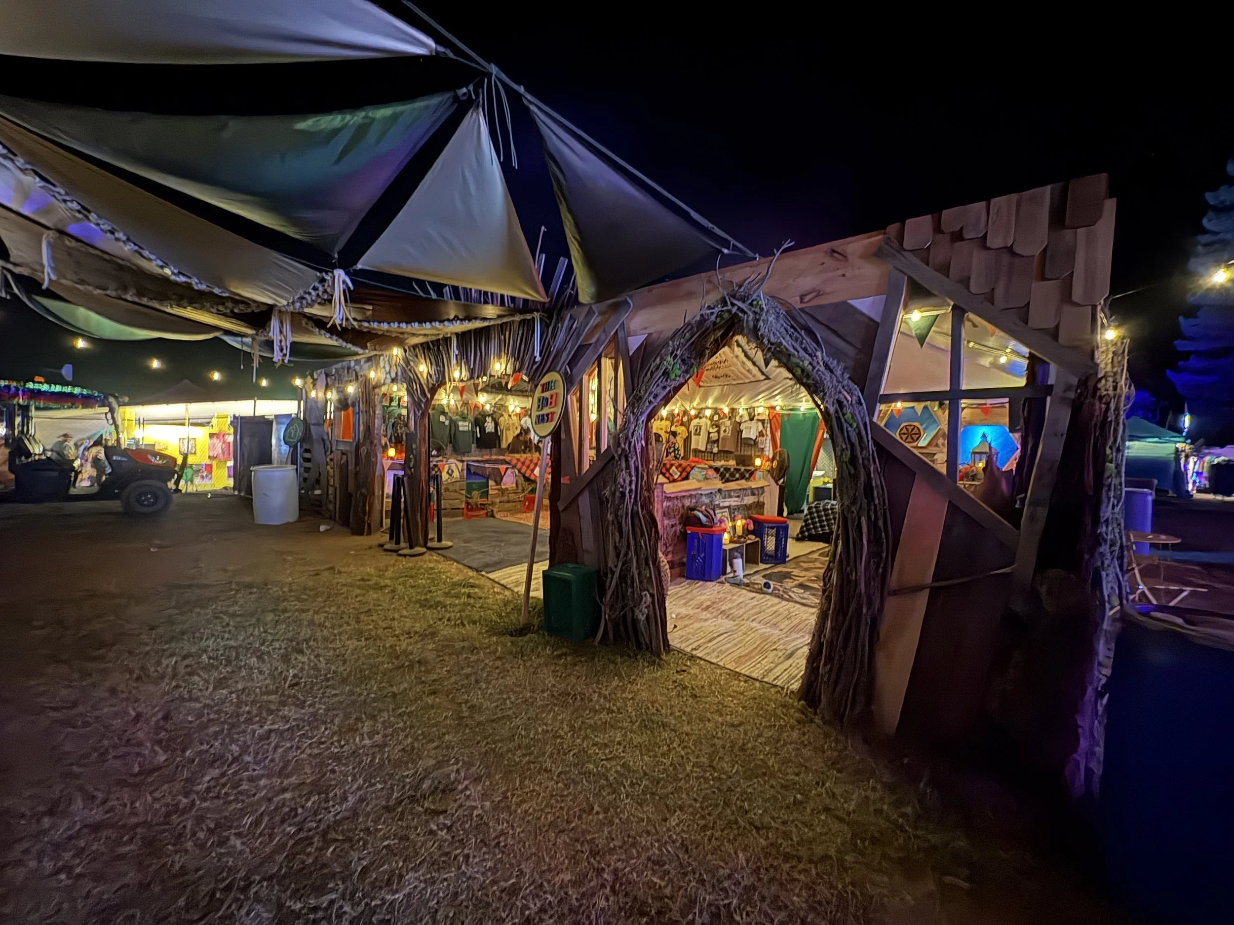Night scene of a decorated outdoor vendor stall with string lights, decorations, and souvenirs. There is a wooden structure with a thatch roof entrance surrounded by trees and open space in the background.