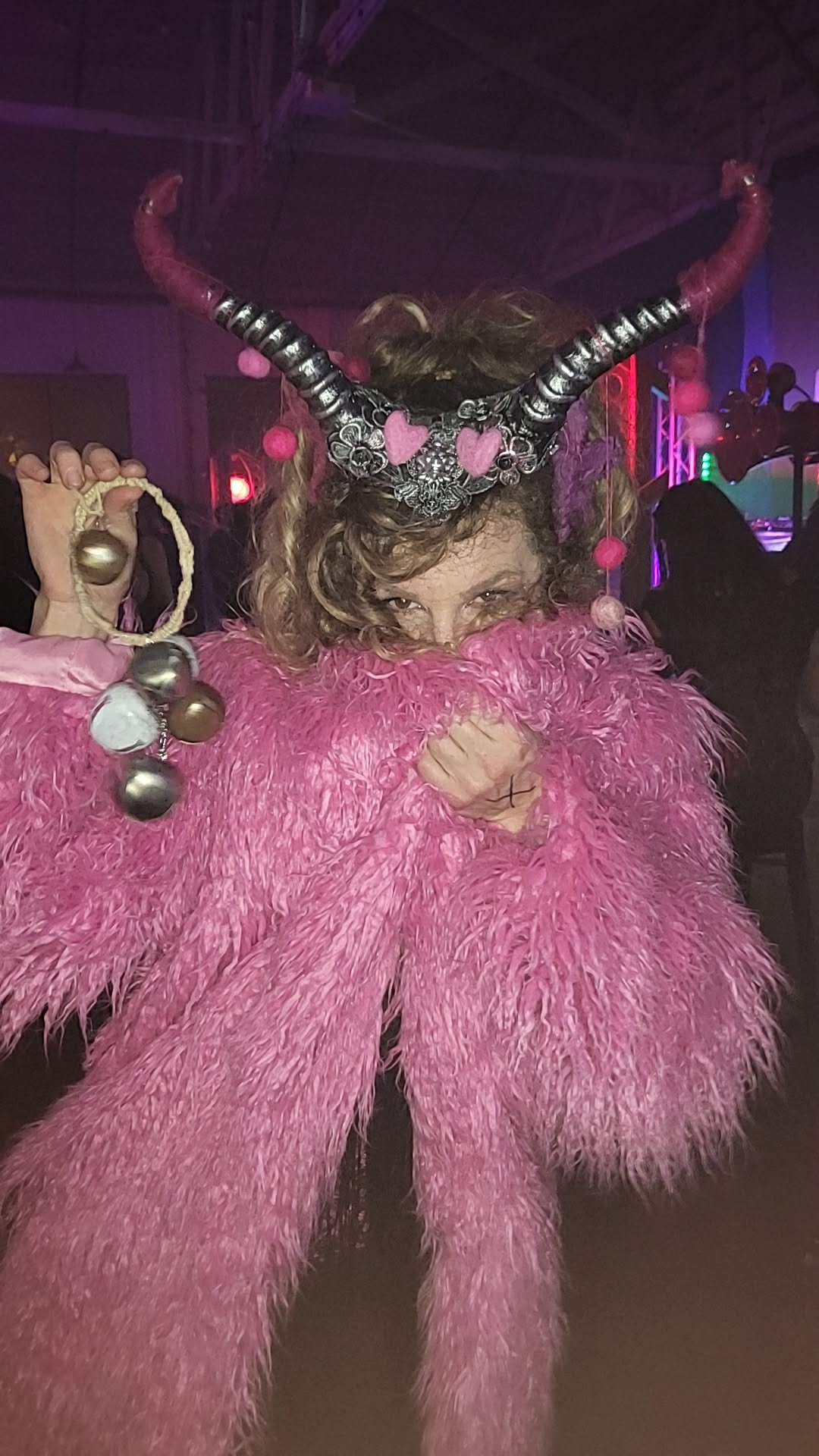 A woman dressed in a bright pink, feathered costume with a headpiece adorned with horns, hearts, and metallic accents. She is holding a jingle bell and smiling at the camera, in a setting with colorful lighting and silhouettes of other people.