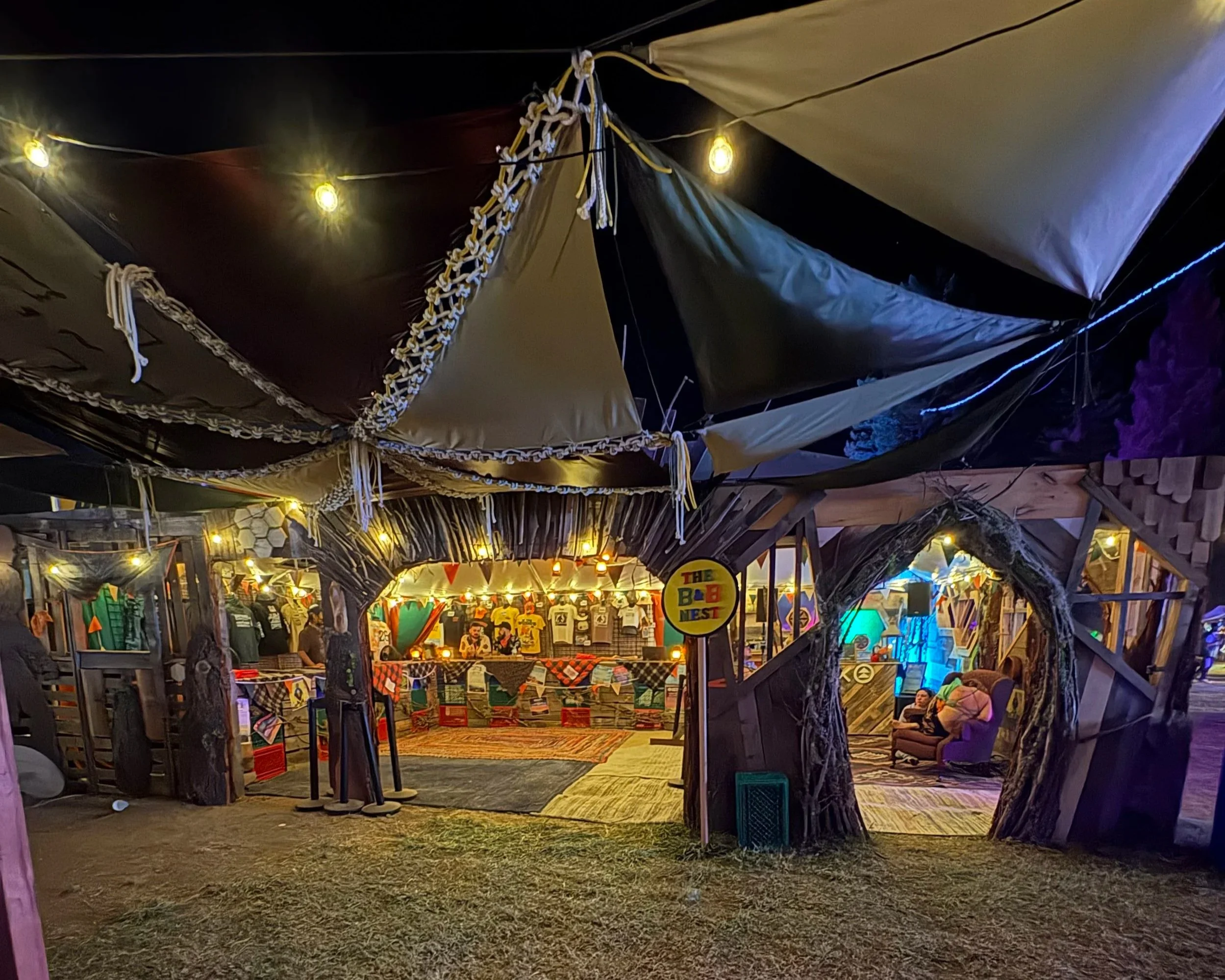 Night scene of a decorated outdoor market or festival with string lights, colorful banners, and various booths selling items. People are sitting and browsing the stalls under a canopy of fabric and lights.