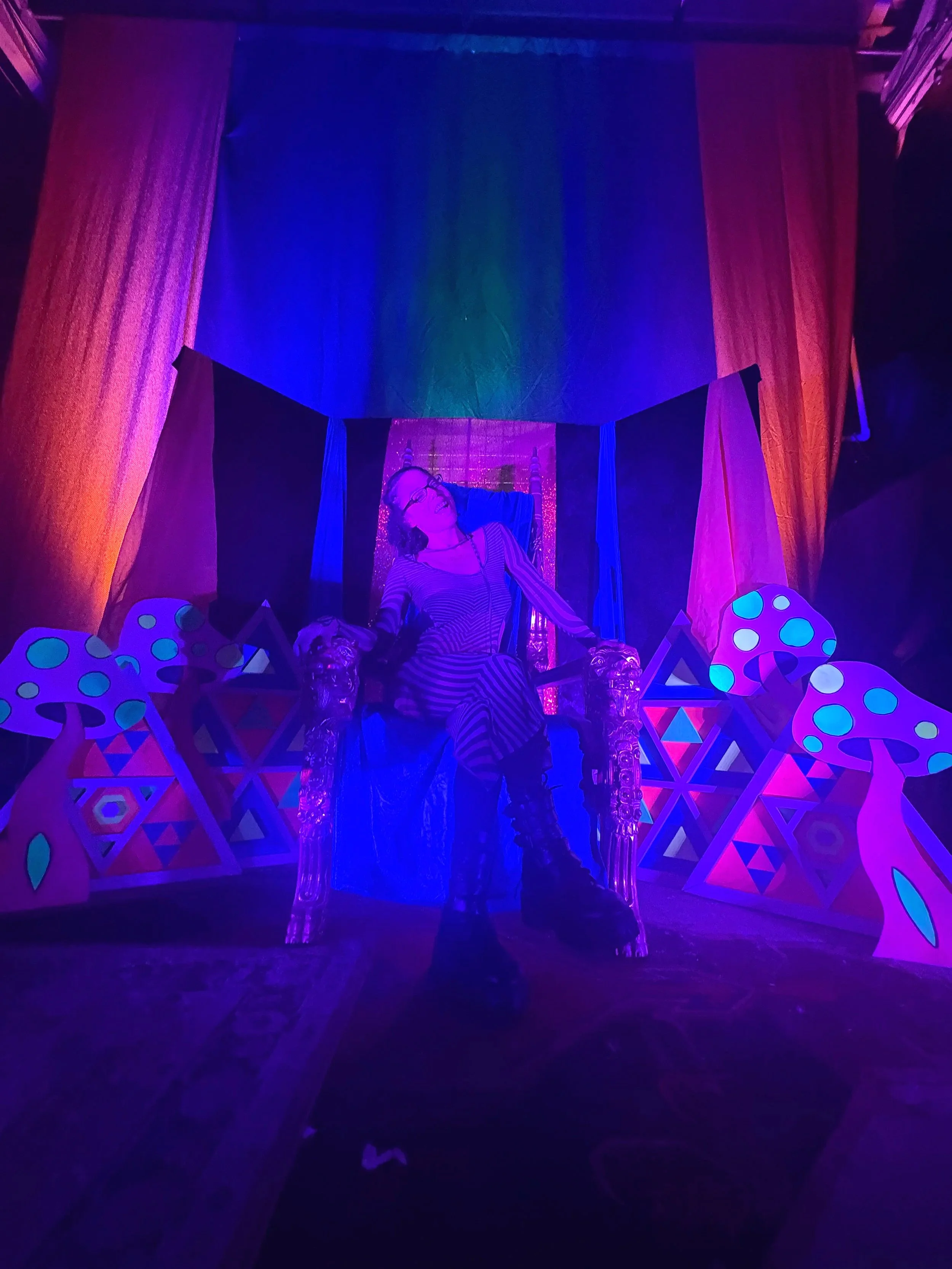 A woman wearing glasses and a striped dress sitting on a perched throne with colorful mushrooms and geometric decorations, illuminated by purple and blue neon lights.