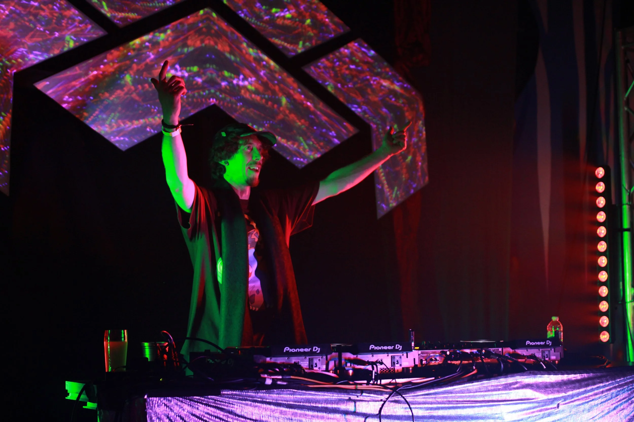 A DJ stands behind a mixing console with Pioneer DJ branding, raising arms, with a colorful, abstract digital display above. The scene is set in a dark environment with vibrant, neon lights.
