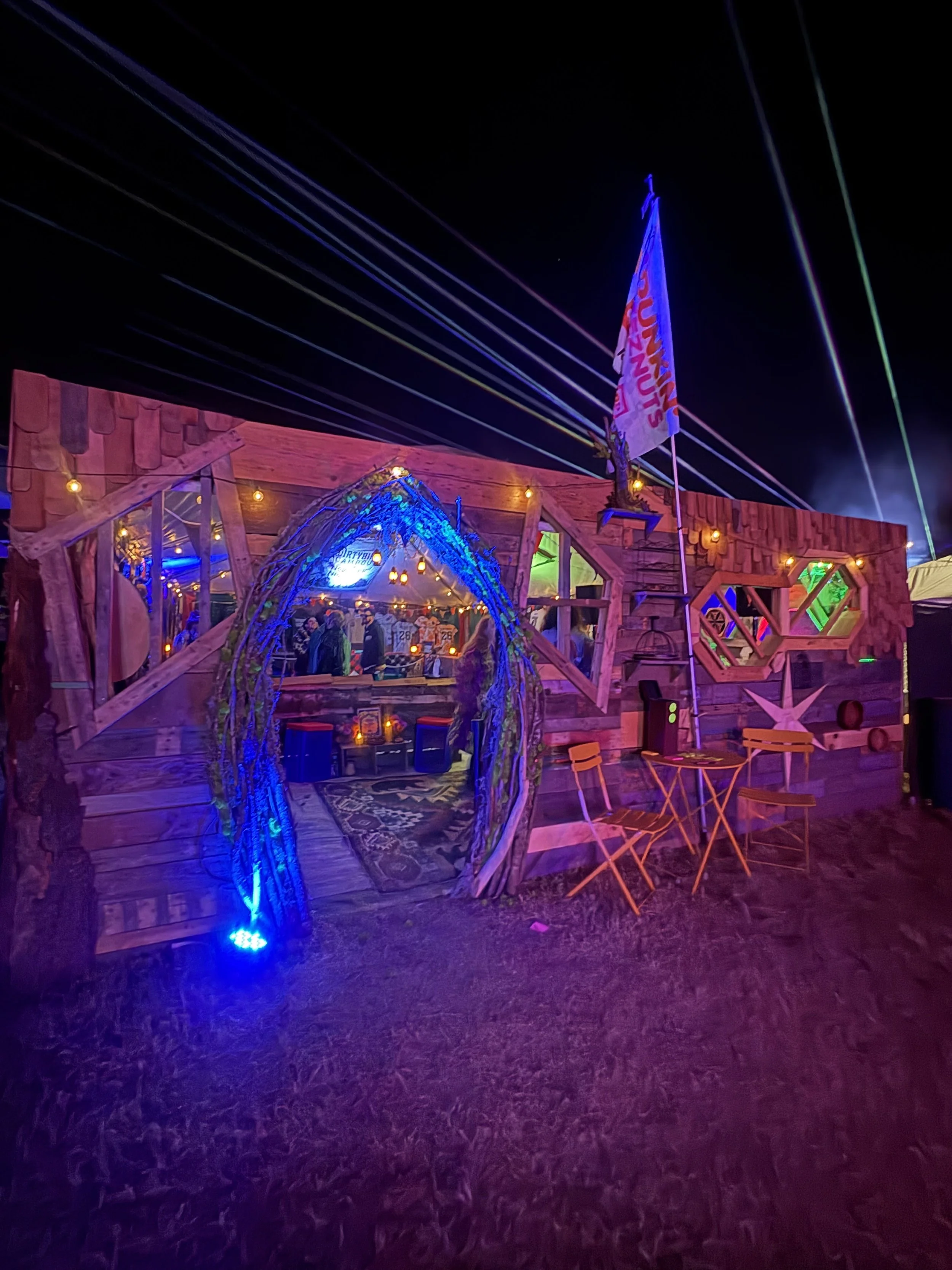 An outdoor fair or festival scene at night with a wooden structure, colorful lights, a flag, and string lights overhead.