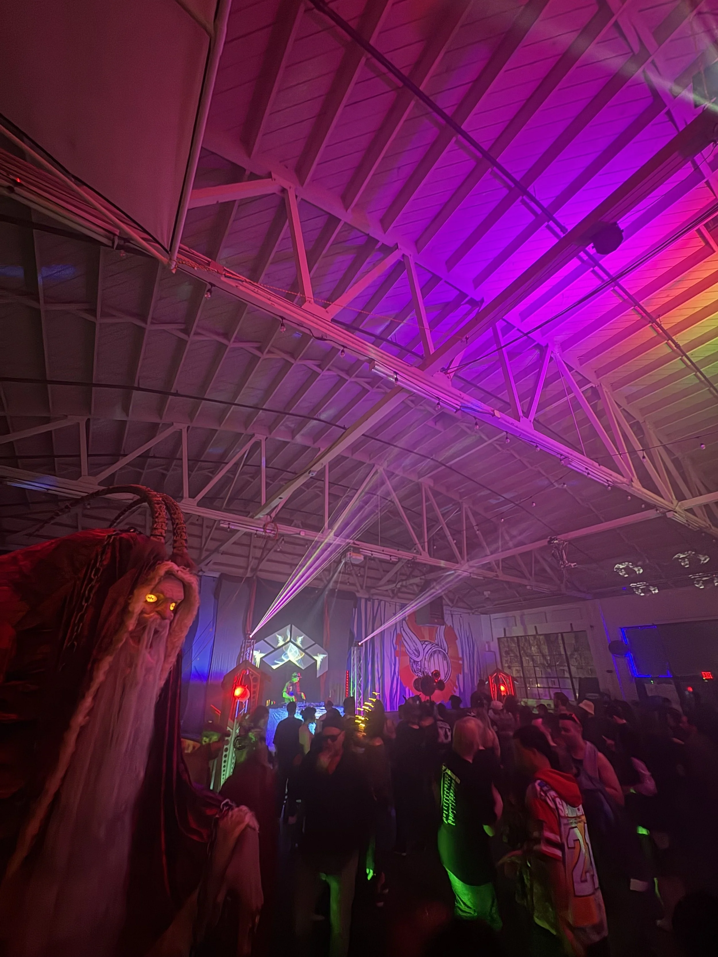 A crowded indoor event with colorful purple, pink, and green lighting, featuring a large crowd of people watching a stage with electronic music and DJ performance.