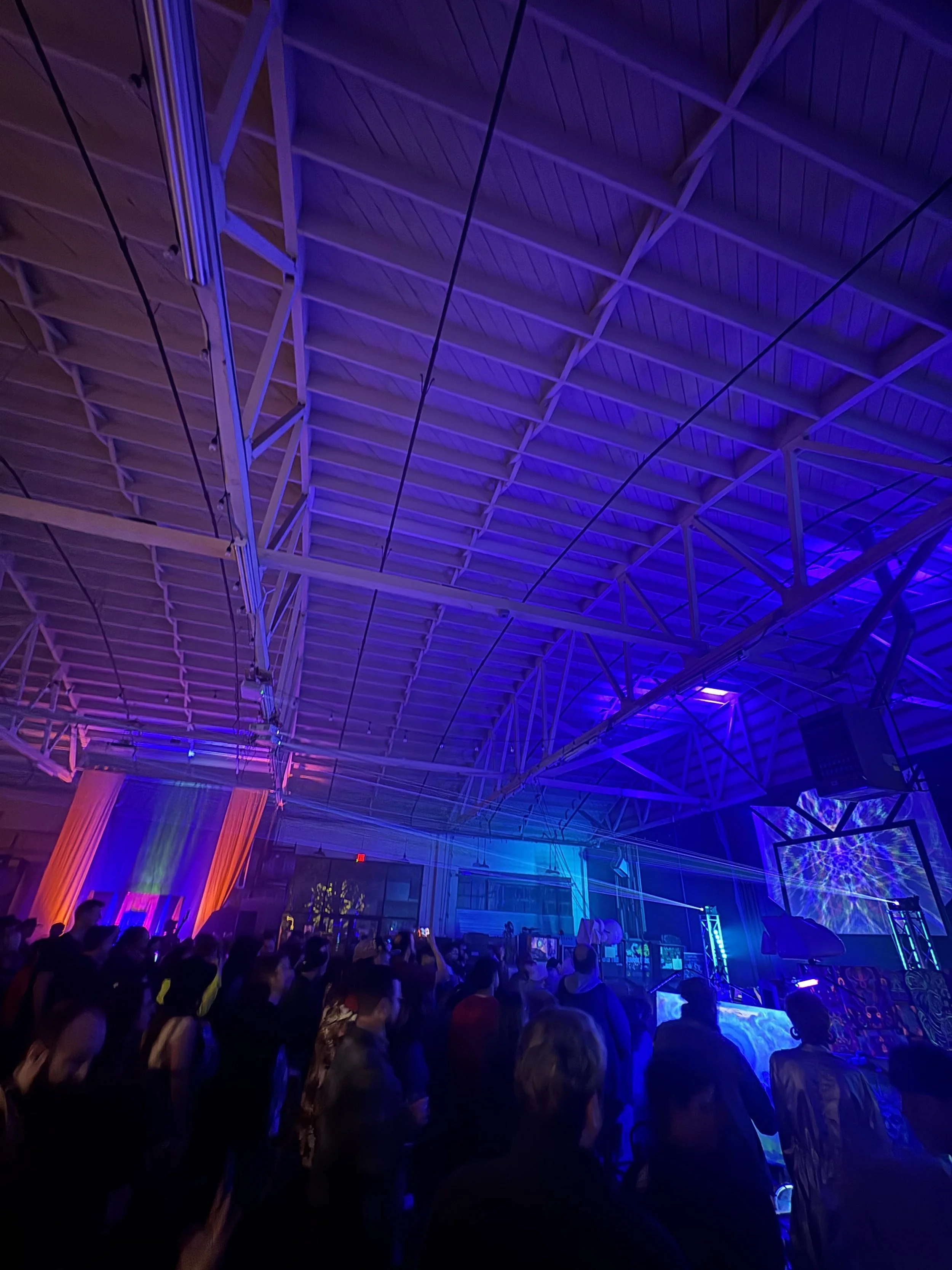 A large crowd of people gathered inside a convention center or warehouse with a high arched ceiling. The space is illuminated with blue and purple stage lighting, and there is a large LED screen displaying vibrant visuals. The attendees are facing a 