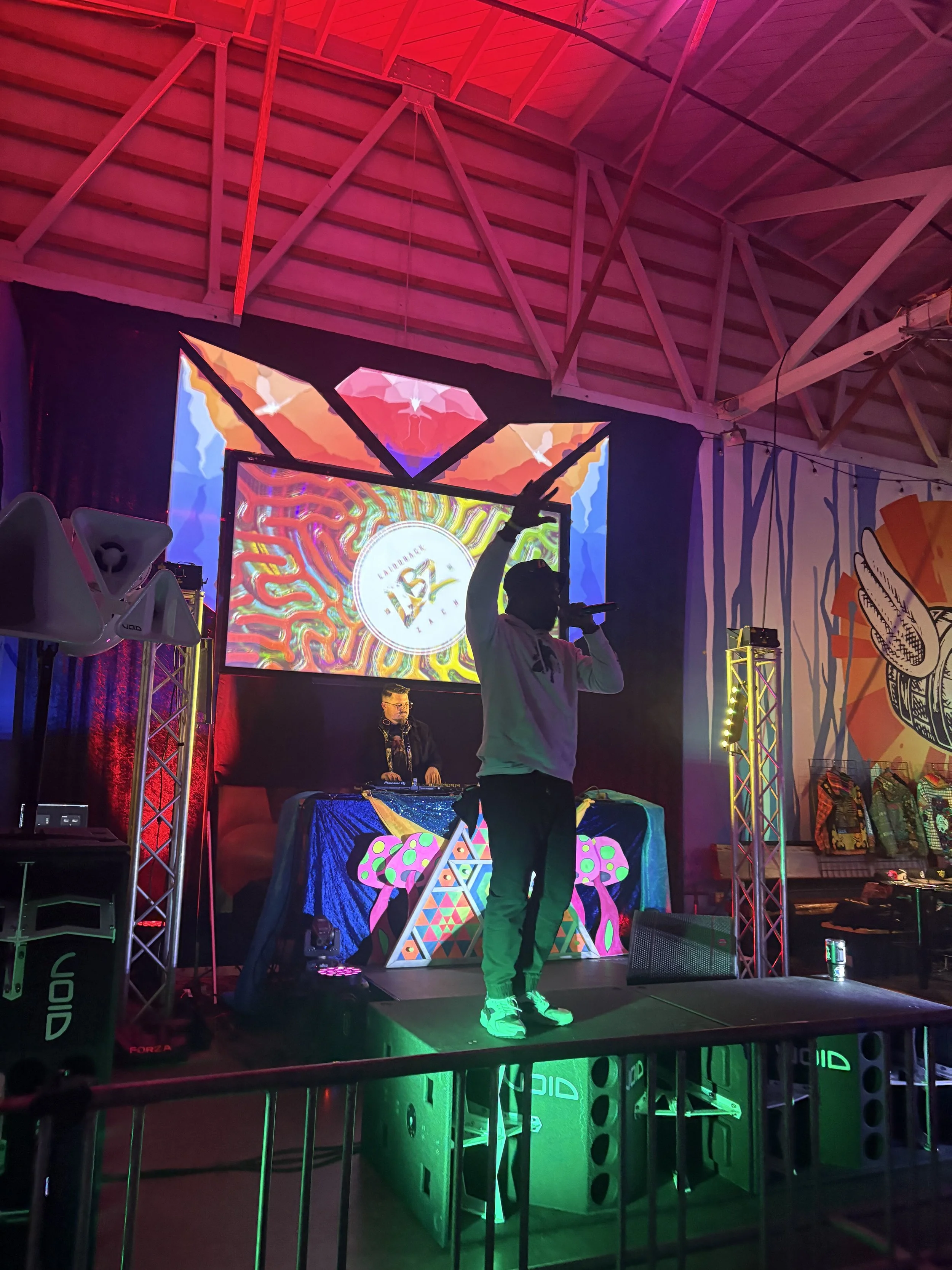 A colorful stage with a DJ performing, a digital screen displaying vibrant artwork and a logo, and a man standing in front of the DJ. The stage is lit with pink, green, and other colorful lights, and there are various decorative and electronic equipm