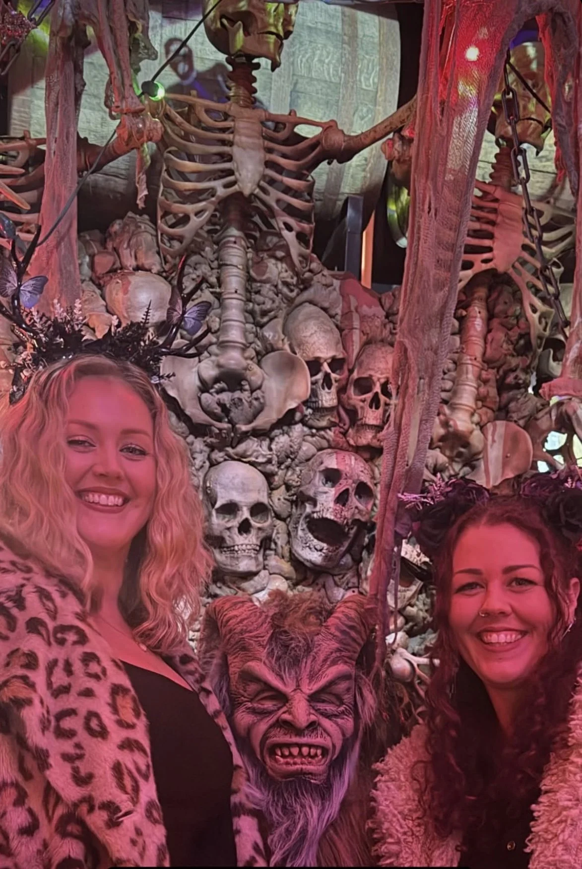 Two women smiling in front of a display of skulls, skeletons, and mythical creature masks at a themed event or exhibition.