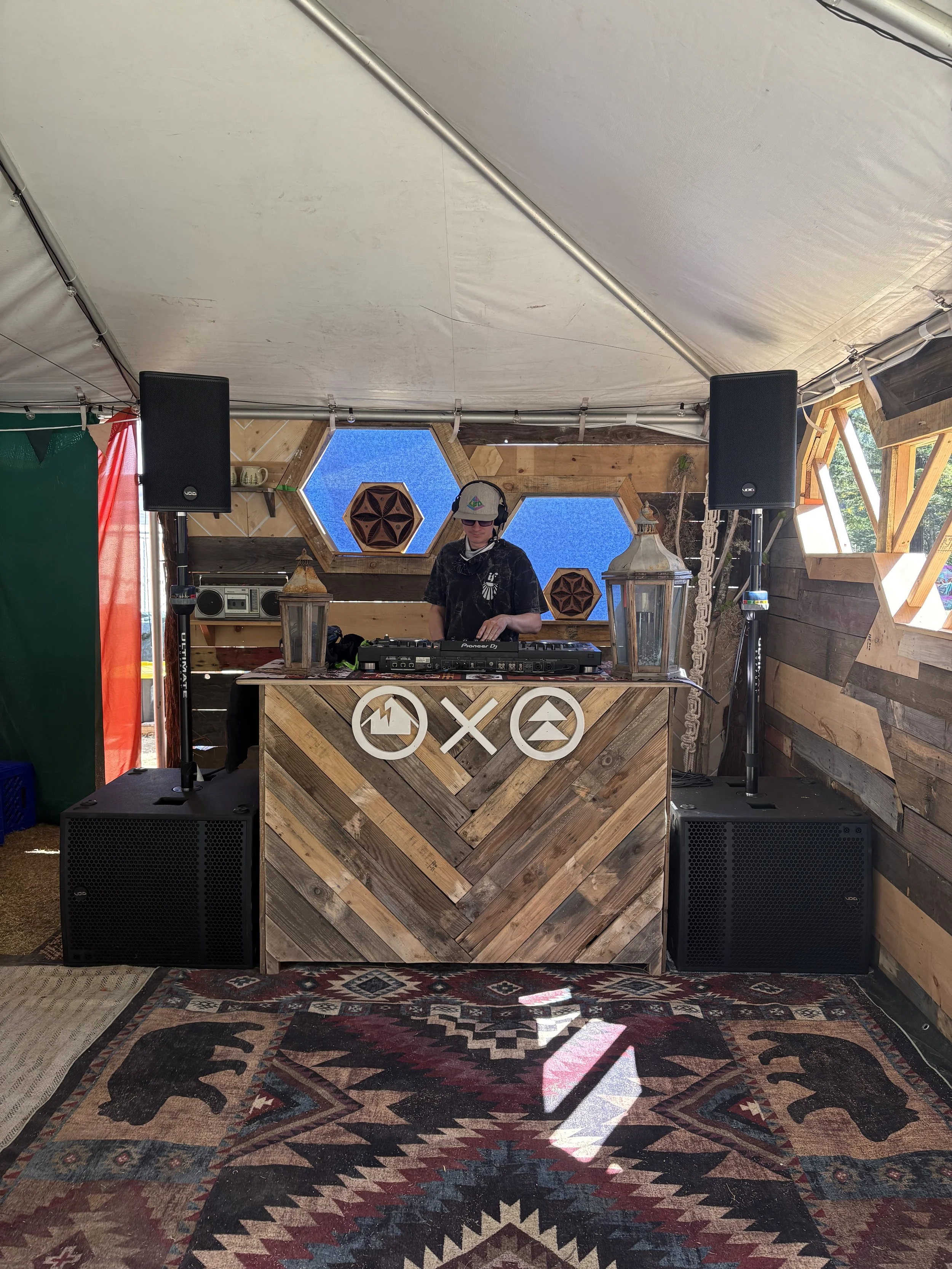 A DJ wearing a cap, sunglasses, and headphones stands behind a wooden DJ booth with two large speakers on either side, inside a tent with wood-paneled walls and hexagonal windows showing a blue sky with clouds. The booth has symbols of a house, a cir