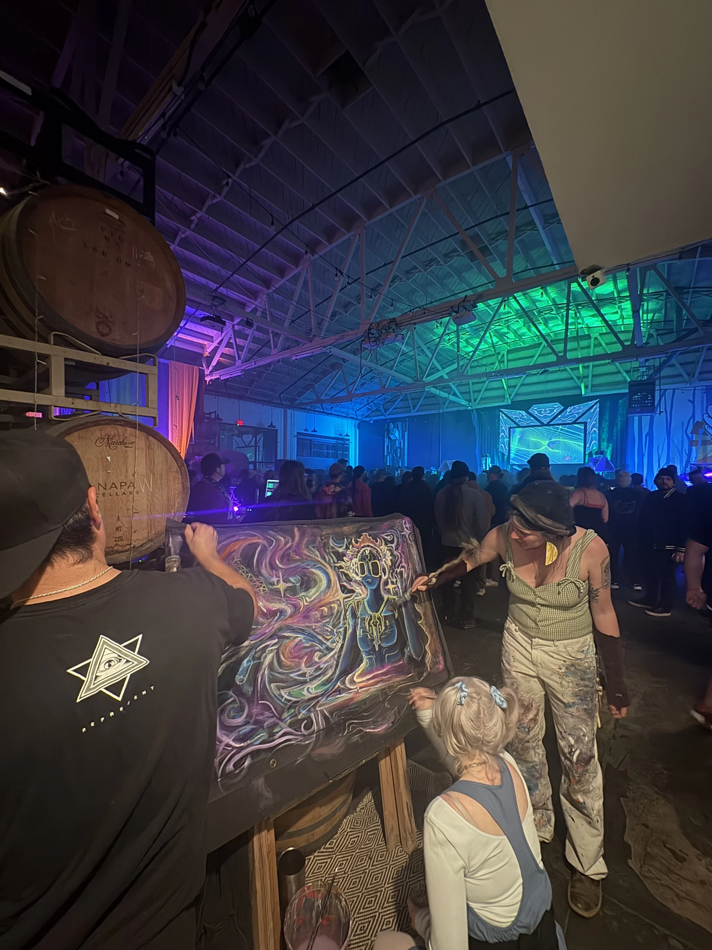 A crowded indoor event with people gathered around a table displaying a colorful, surreal artwork. The background has a large projection screen and vibrant purple and green lighting on the ceiling. Some people are standing and observing, while a woma