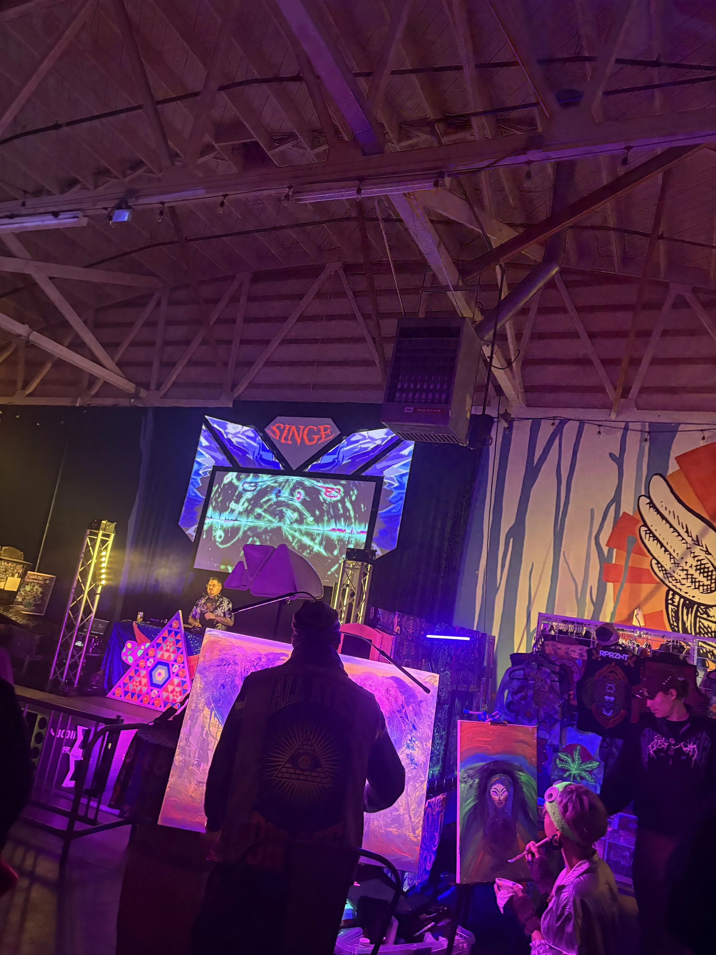 An indoor art event or exhibition with colorful, fluorescent paintings on display. Several people observe the artwork, and there is a large screen displaying digital visuals on the wall. The ceiling features exposed wooden beams, and a large speaker 