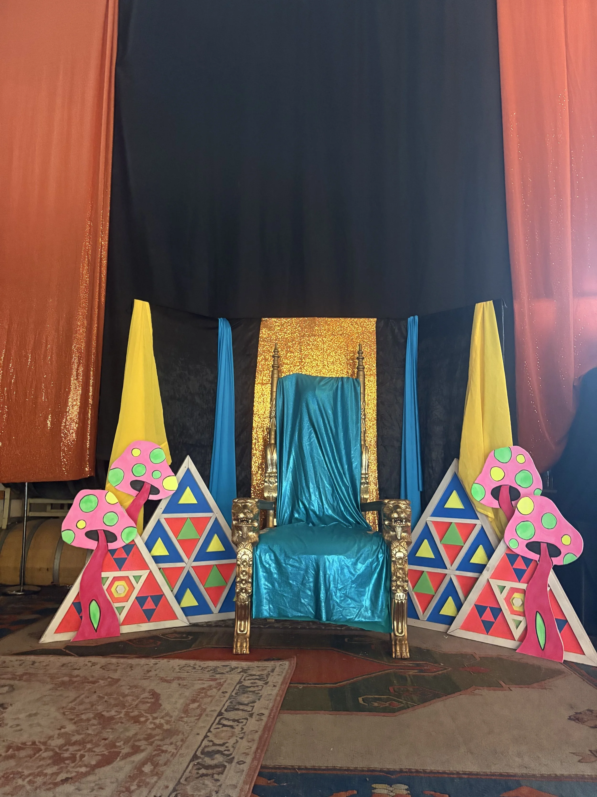 Decorative throne with a blue velvet fabric and gold ornate arms, set against a gold glittery backdrop, flanked by colorful geometric painted cardboard cutouts of mushrooms and triangles, and red, yellow, and blue drapes.