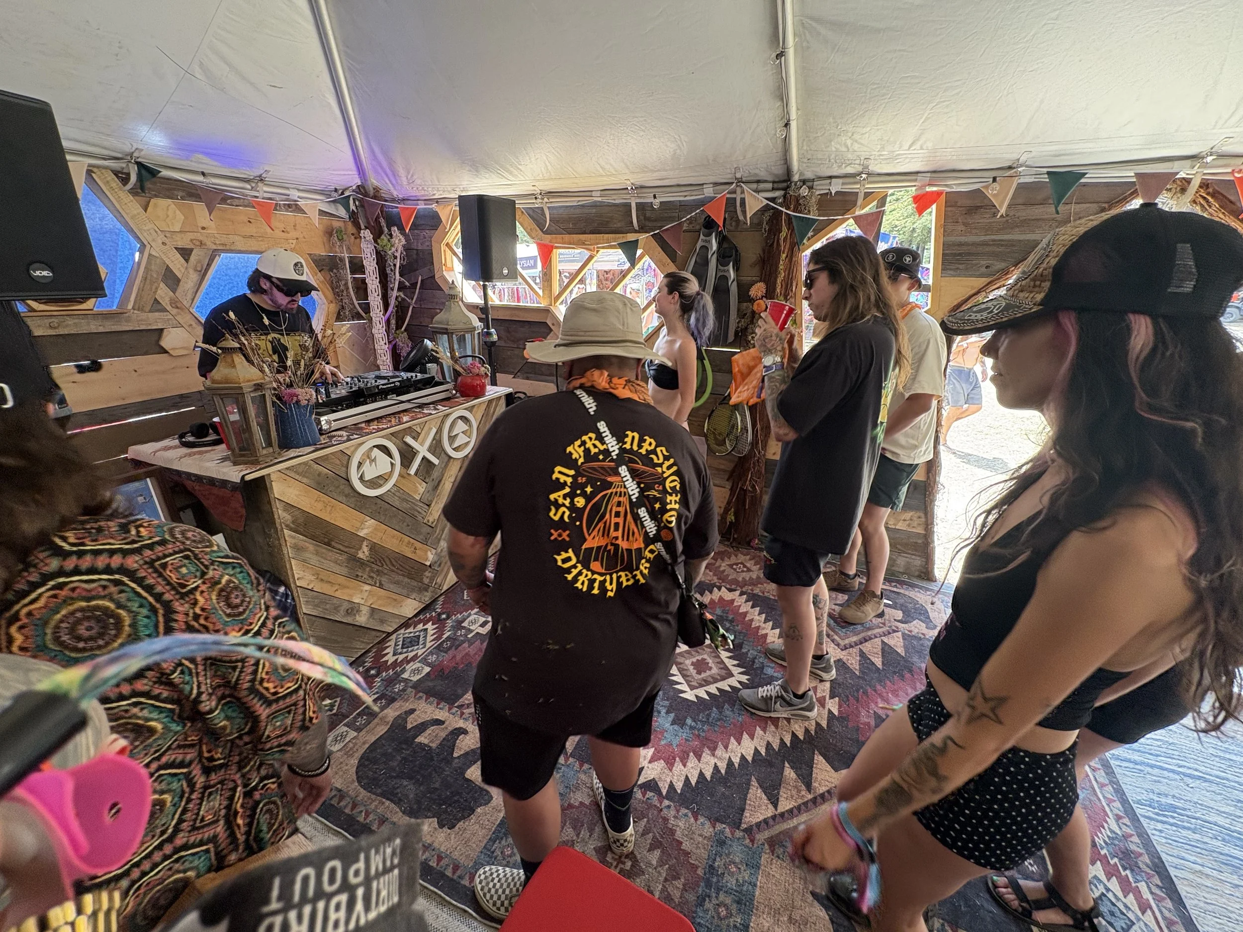 A group of people gathered around a DJ booth at an indoor event, with colorful bunting hanging from the ceiling and various snacks and items on the table.