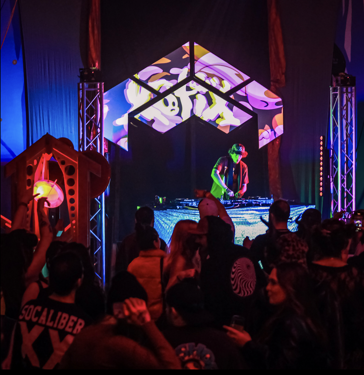 A DJ performs on stage at a concert with colorful lighting and digital screens displaying abstract artwork. The audience watches and takes photos.