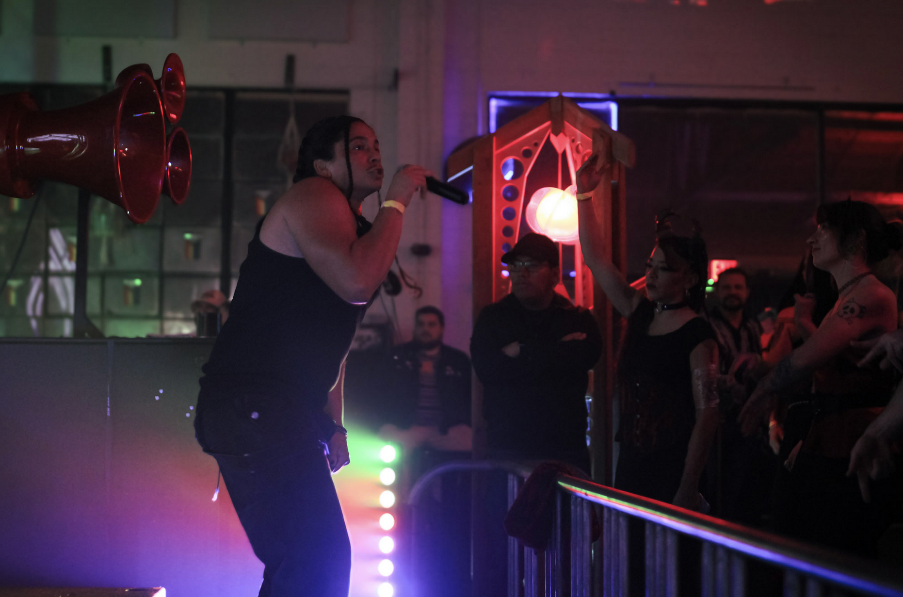 A person with long dark hair singing into a black microphone on a stage with colorful lighting, in front of an audience in a dark room with neon lighting and loudspeakers. illuminous events llc