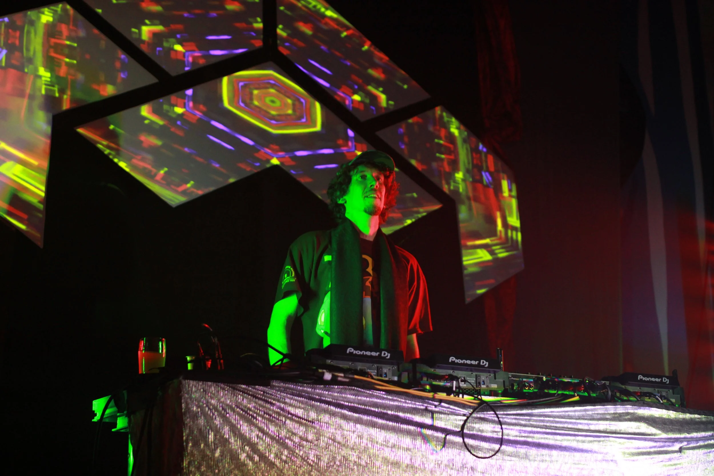 A DJ with curly hair in a hooded jacket performing at a club with colorful, neon digital visuals projected above and behind them.