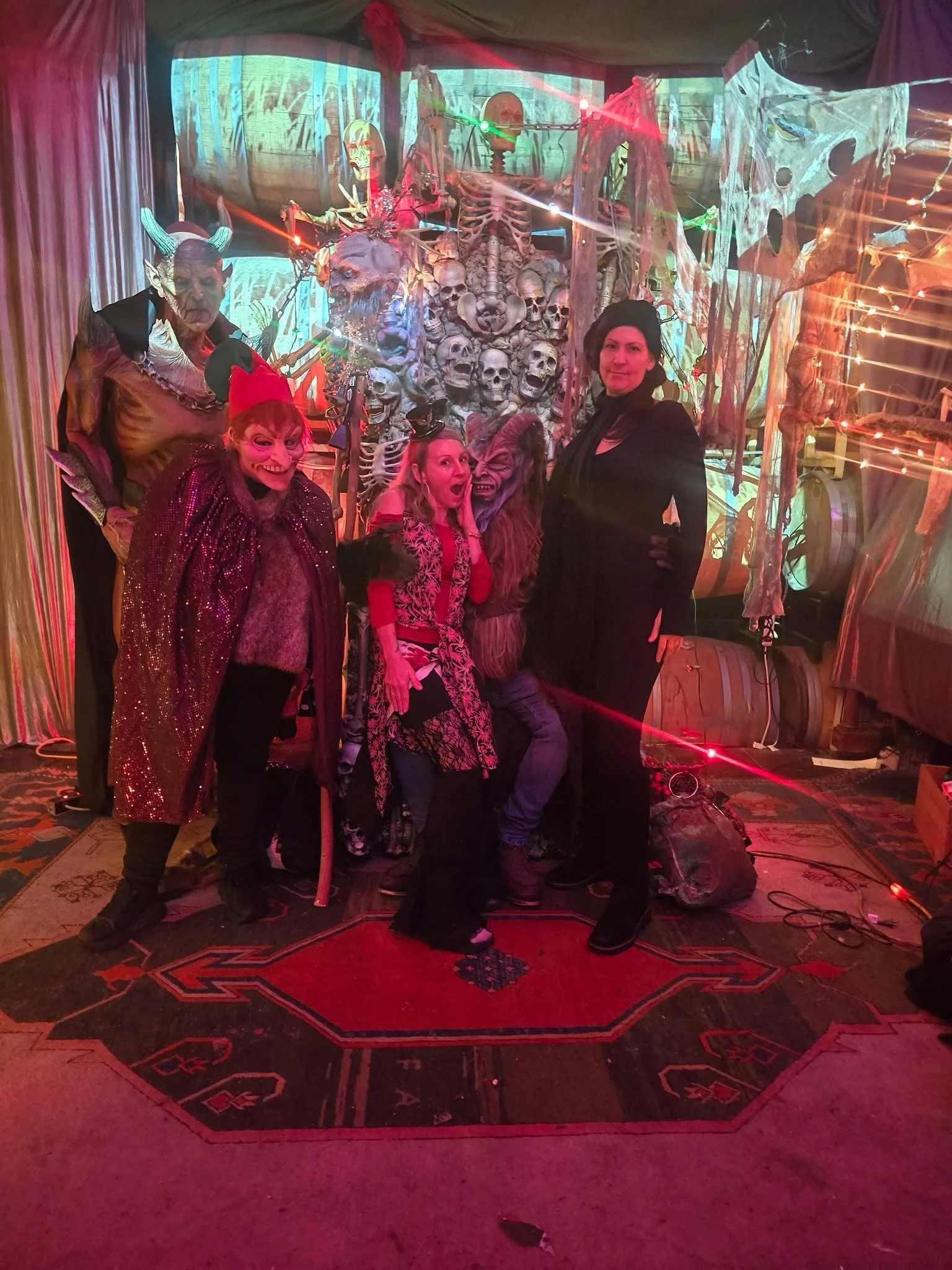 Four people in Halloween costumes posing in front of a spooky backdrop with skulls, skeletons, and cobwebs, illuminated by red and green lights.
