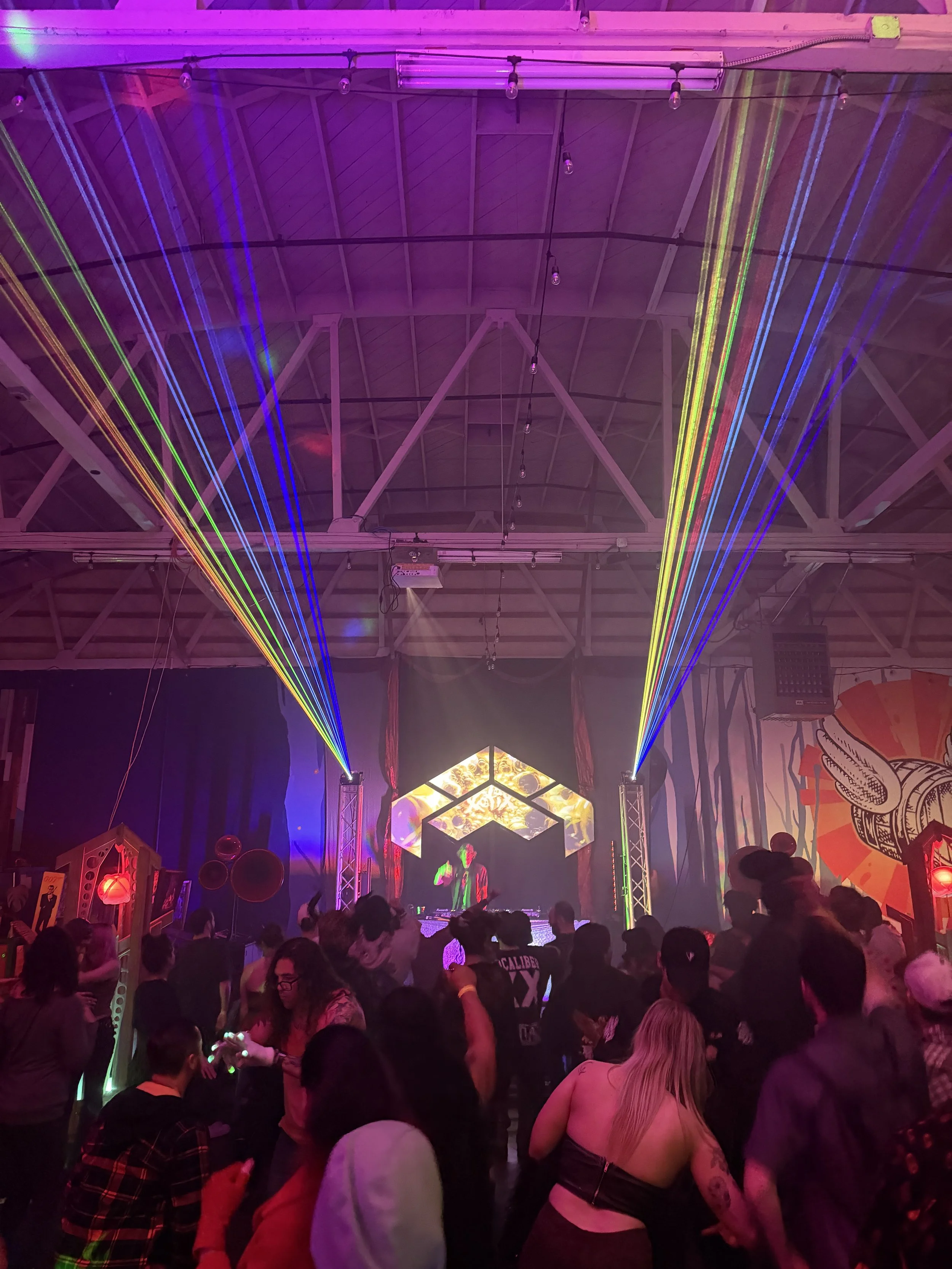 A crowded indoor club scene with colorful laser lights and a DJ performing on stage. People are dancing and enjoying the music.