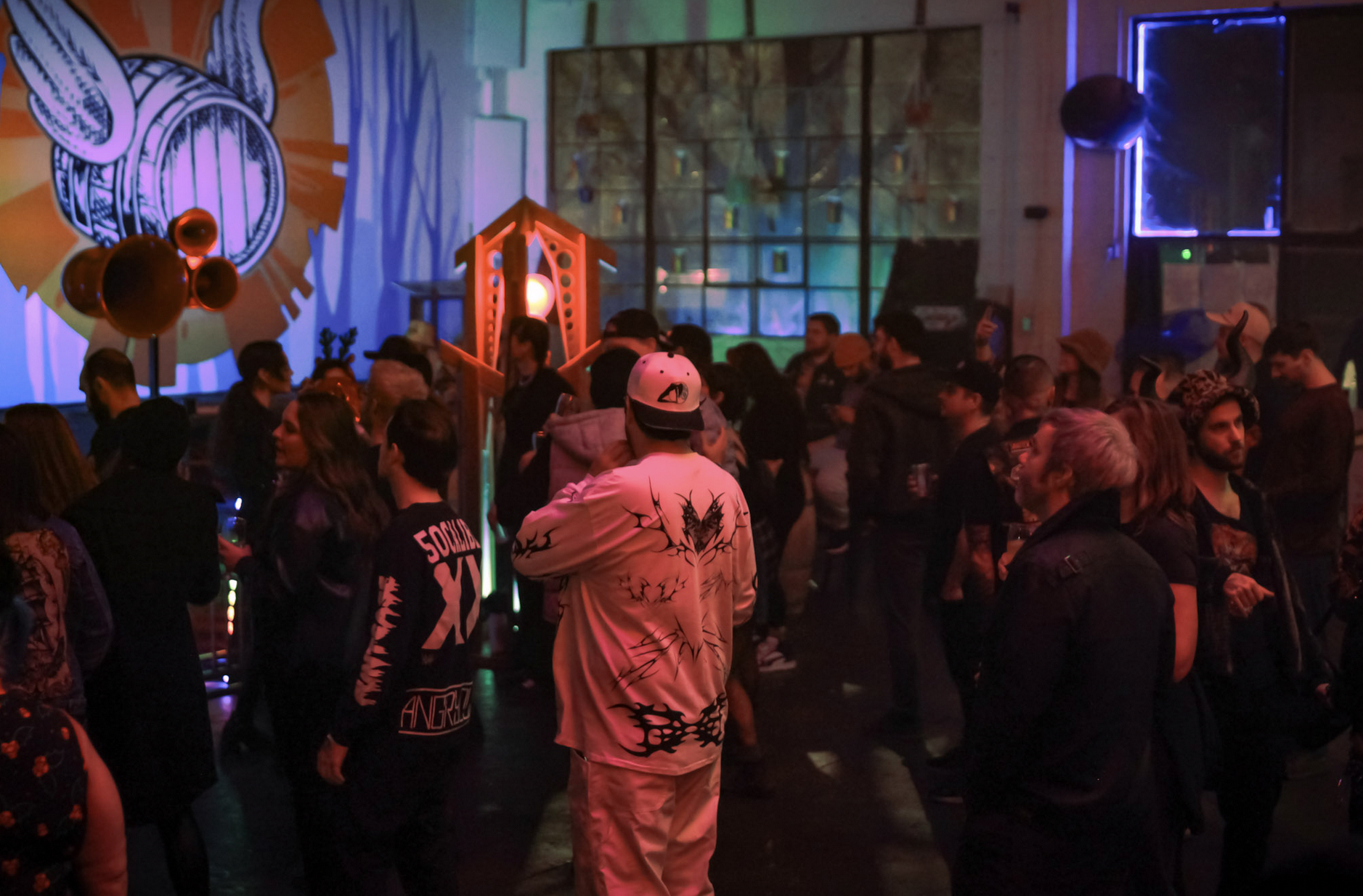 People at a crowded indoor music event with colorful lighting, a large mural on the wall, and various decorations. illuminous events llc