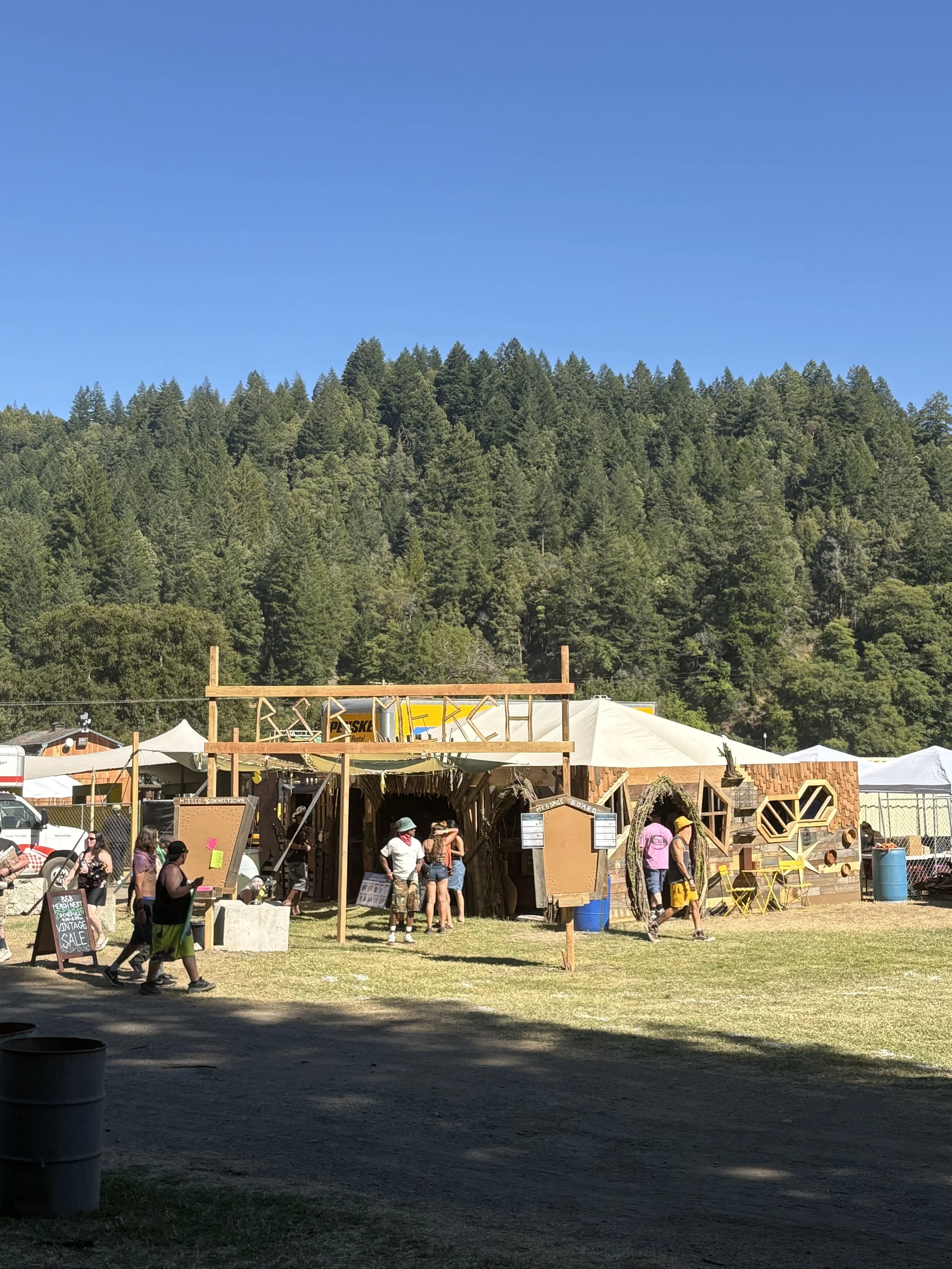 People visiting a wooden booth at an outdoor event with tents, surrounded by trees and a mountain backdrop.
