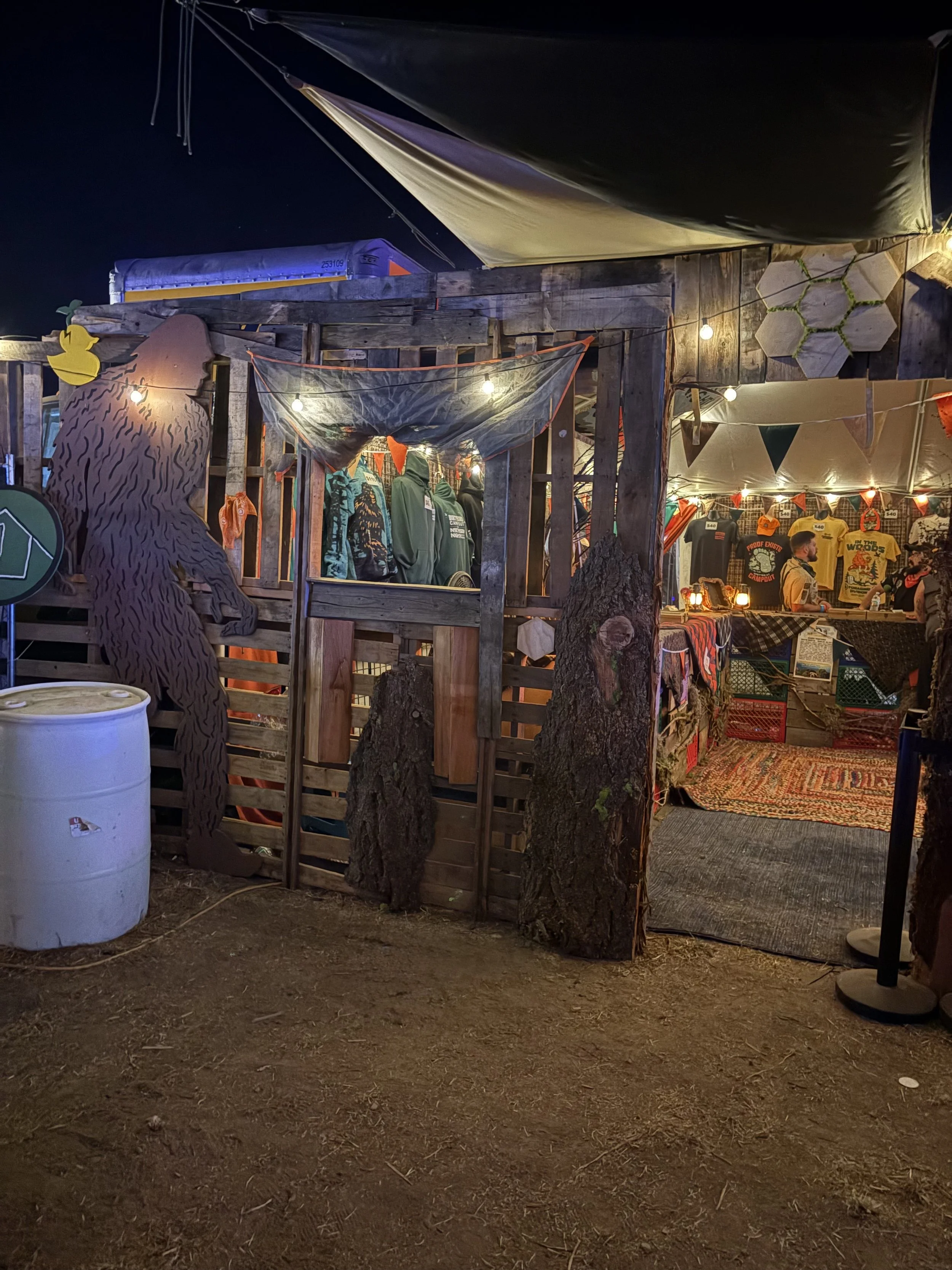 Nighttime scene of a rustic outdoor market stall decorated with string lights, displaying clothing items, with a wooden structure, trees, and a textured rug on the ground.