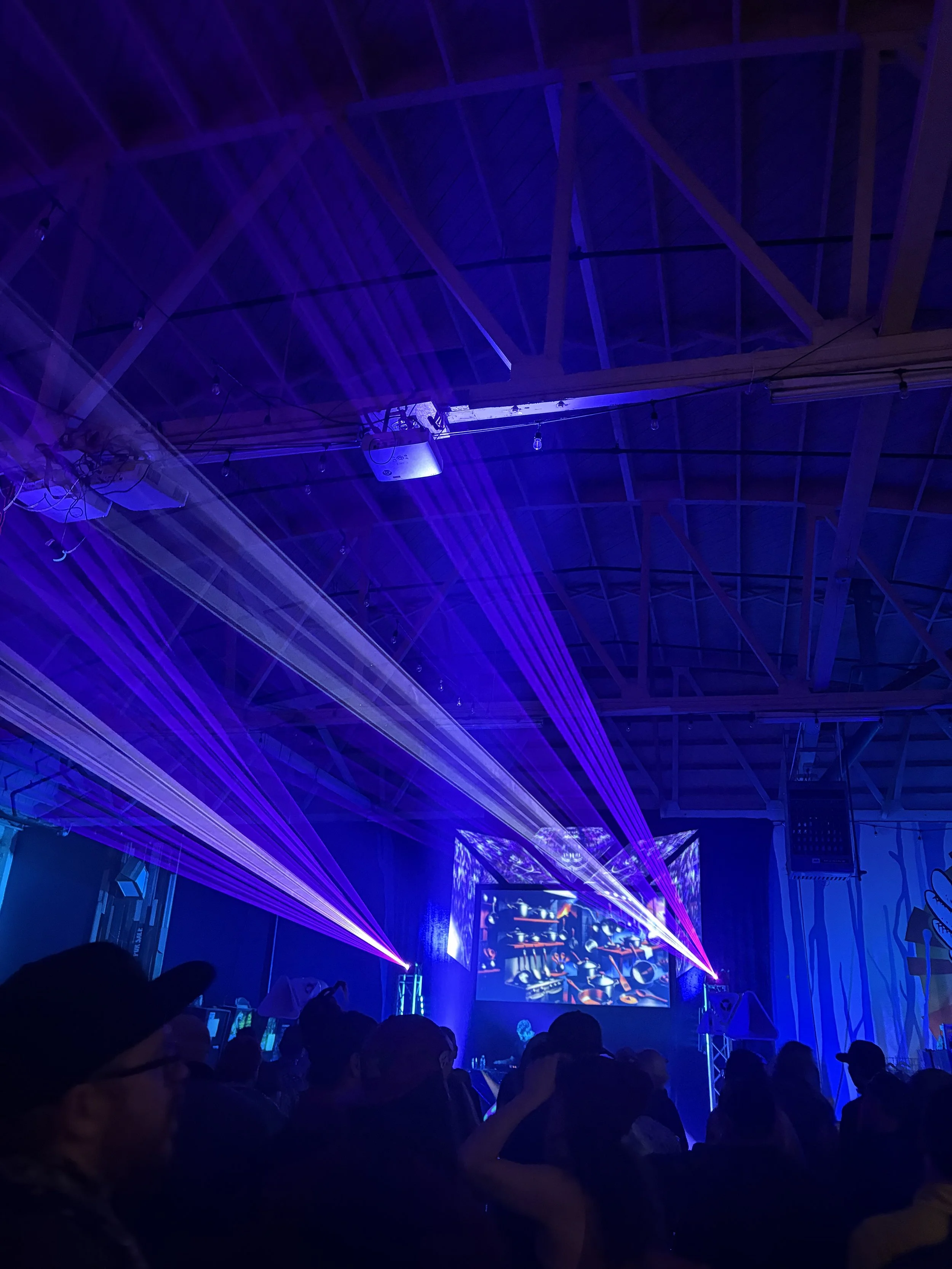 A dark indoor concert or event space with blue and purple stage lighting, laser lights projecting across the ceiling, and a crowd of people watching a performance on a large screen.