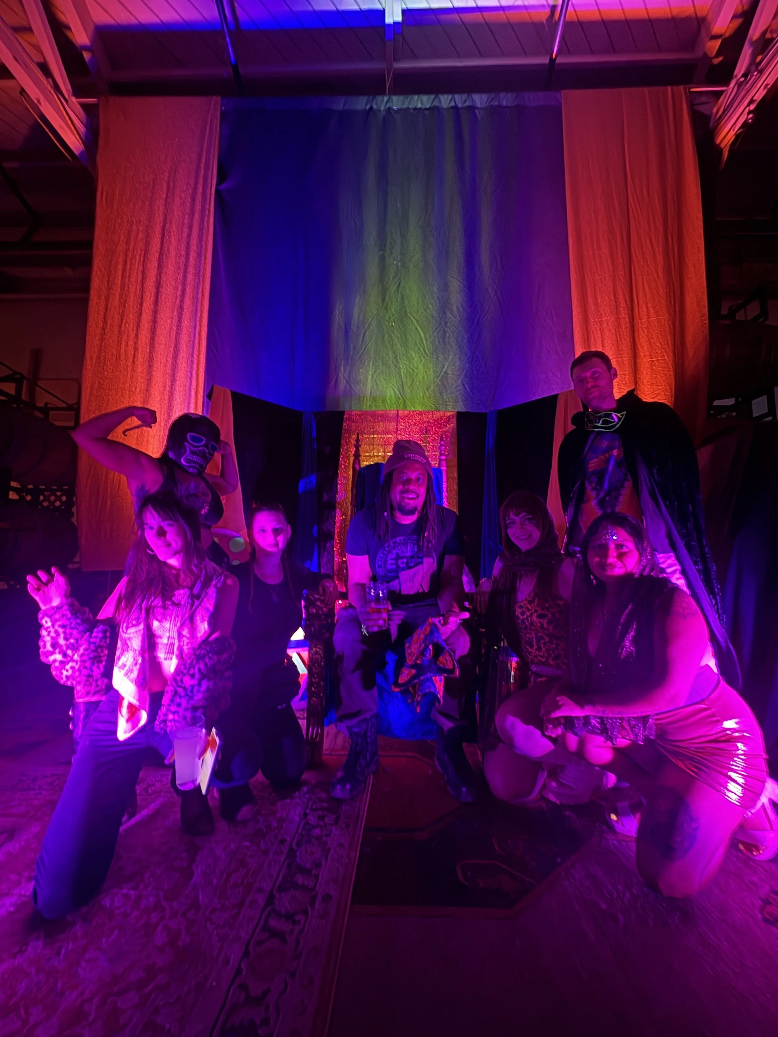 A group of people, including children and adults, posing for a photo under colorful, neon lighting with red, purple, and green hues, in a room with a decorated curtain backdrop.