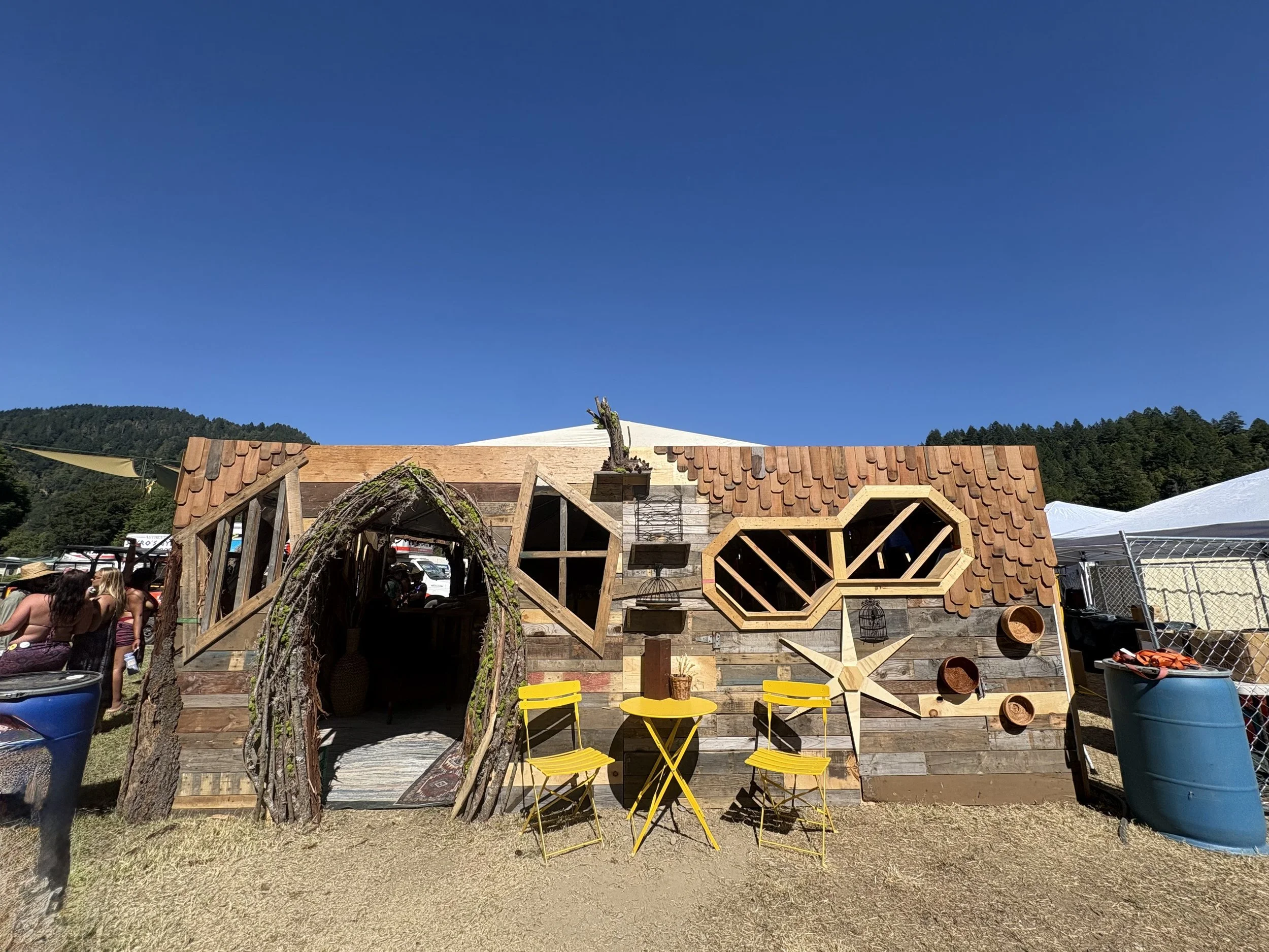 A scene of a construction or outdoor setup with a partially built wooden structure, bright yellow chairs and tables, a decorative piece resembling a tree, and various building materials, against a backdrop of mountains and a clear blue sky.