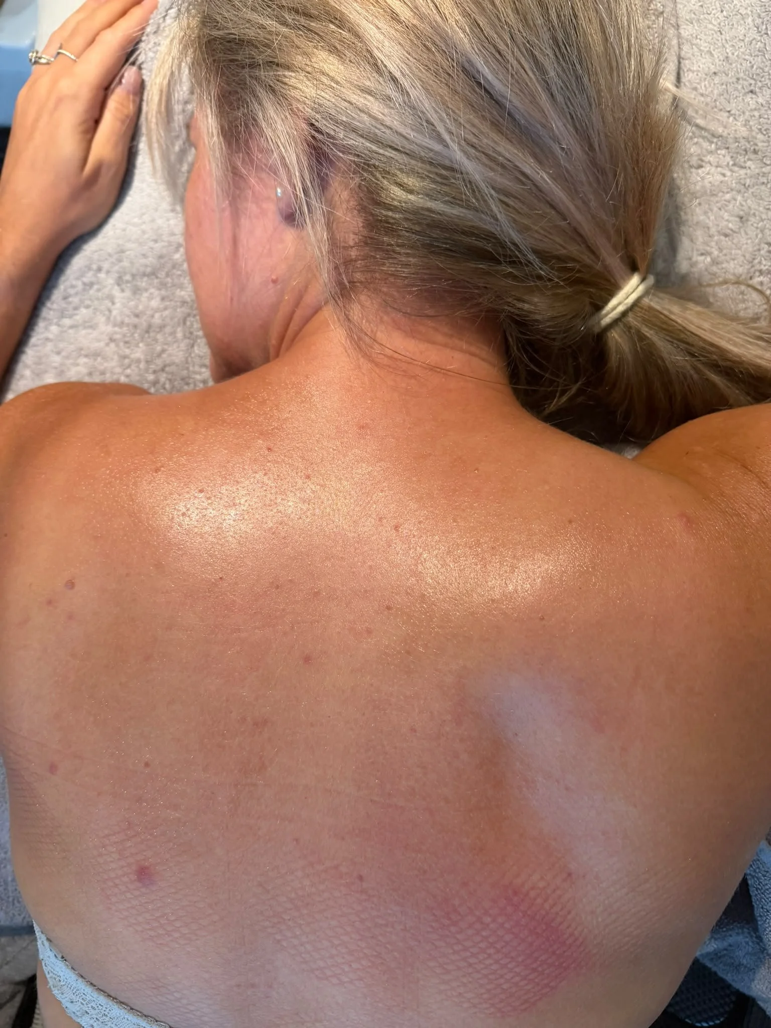 Close-up of a person's back, shoulder, and neck, with the person's head resting on the ground. The individual has blonde hair tied back with a hairband, and the skin appears glistening, possibly due to sweat or lotion.