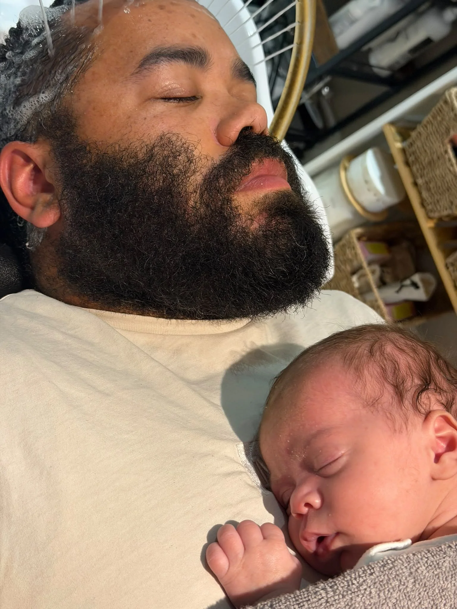 A man with a thick beard and closed eyes is lying down with a baby sleeping beside him, both resting on a light-colored surface.