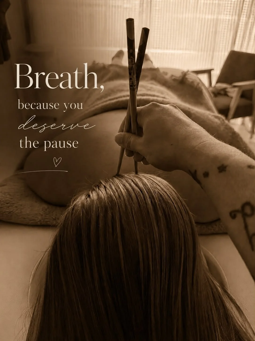 A person lying on a massage table with their back exposed, holding incense sticks over their head. The background shows a relaxed indoor setting with a chair and curtains. An overlay text reads: "Breath, because you deserve the pause."