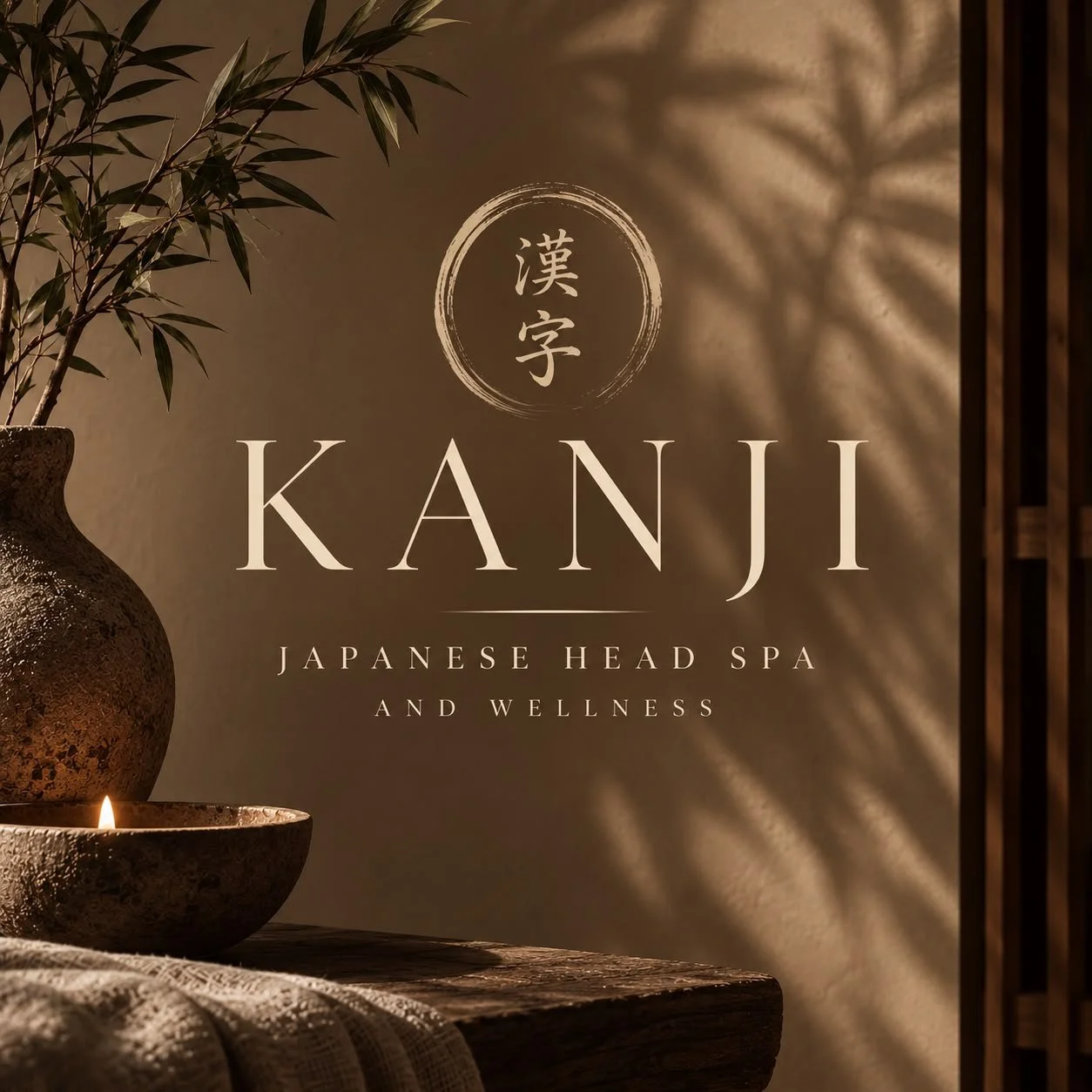 Decorative scene promoting KANJI Japanese head spa and wellness center, featuring a large brown vase with green plant leaves, a small candle, and a textured cloth on a wooden surface, with ambient shadows and warm lighting.